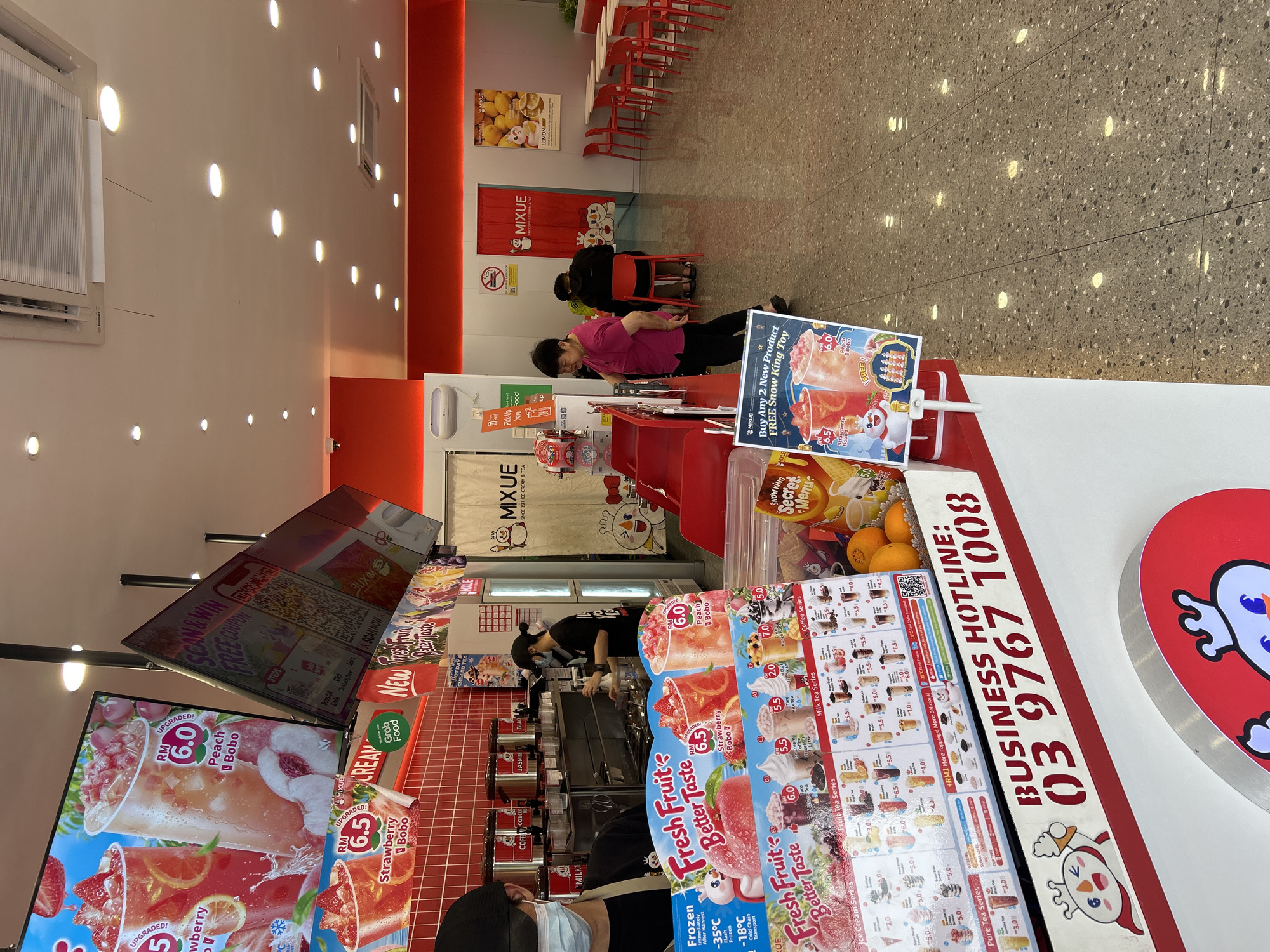 The image displays the interior of a brightly lit Mixue ice cream and tea shop, consistent with a commercial establishment in Subang Jaya City Council, Malaysia, given the "03" area code in the visible hotline number. At the counter, two staff members, wearing masks, are actively preparing beverages. Two customers are present near the counter: one in a pink shirt is standing, and another is partially visible in the foreground. In the background, red chairs and tables are arranged, with an additional customer seated.
The shop prominently features large digital and printed menu displays advertising a variety of drinks. Notable items include "Strawberry Bobo" (RM 6.5) and "Peach Bobo" (RM 6.0), alongside categories such as "Ice Cream Series," "Fruit Tea Series," "Milk Tea Series," "Coffee Series," and "Pure Tea Series." Promotional materials announce a "Buy Any 2 New Product FREE Snow King Toy" offer and encourage customers to "SCAN & WIN FREE COUPON." The shop also advertises "Grab Food" for delivery or pickup. Visible text identifies the brand as "MIXUE SINCE 1997 ICE CREAM & TEA" and provides a "BUSINESS HOTLINE: 03 9767 1008." Other details include information about frozen ingredients ("-35°C Flash Frozen -18°C Cold Chain Transport"), sugar level options, and a no-smoking sign. The setting features a tiled floor with recessed lighting, creating a clean and modern atmosphere.