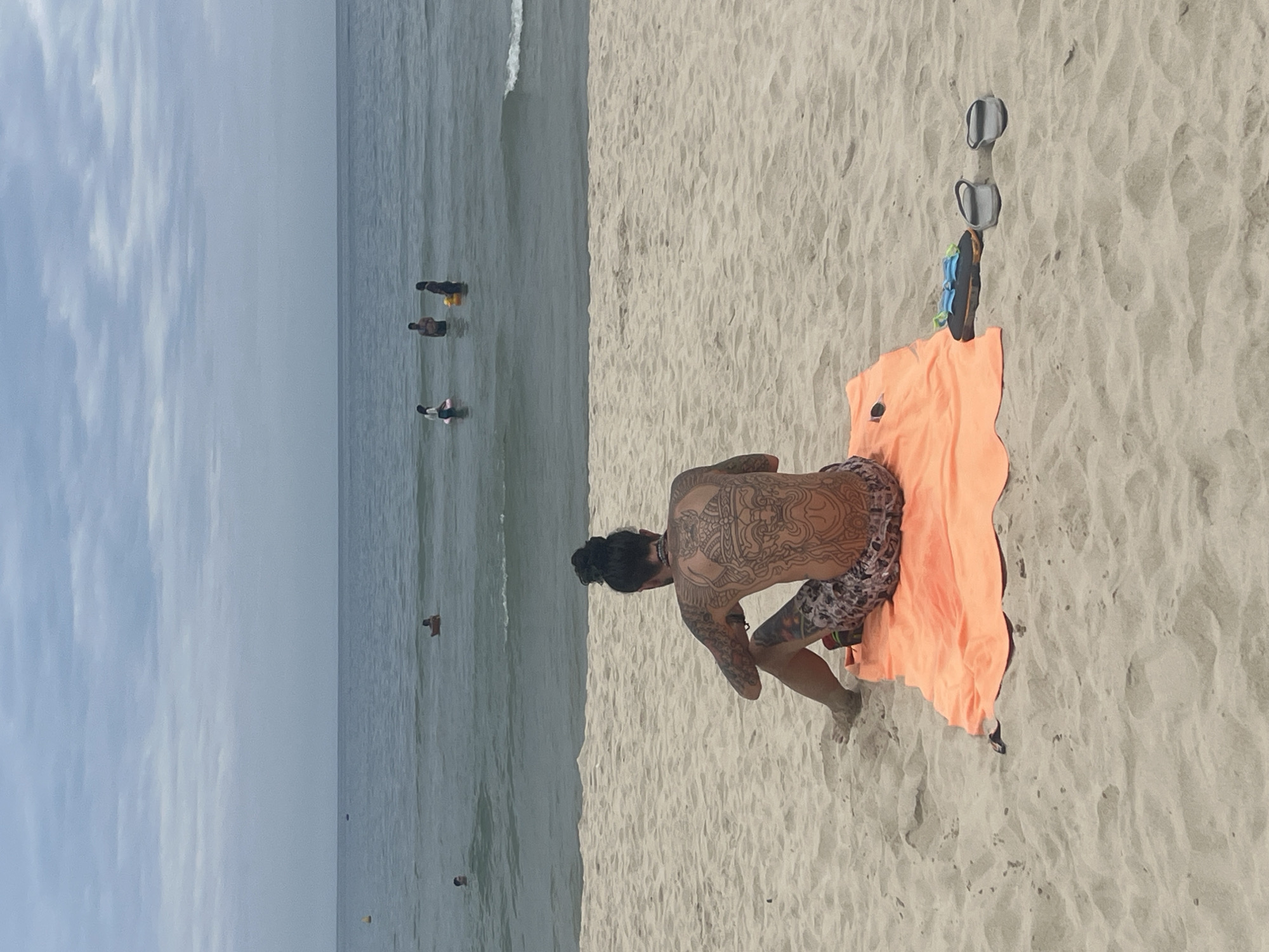A man with extensive back tattoos sits on an orange towel on a sandy beach, facing the ocean. He wears patterned swim trunks. In the background, several people are visible in the water, some swimming and others standing. The sky is overcast, with a light gray haze. Two pairs of flip-flops and a pair of goggles are placed on the sand near the towel. This scene is consistent with a beach in Đà Nẵng, Vietnam, a coastal city known for its sandy shores.