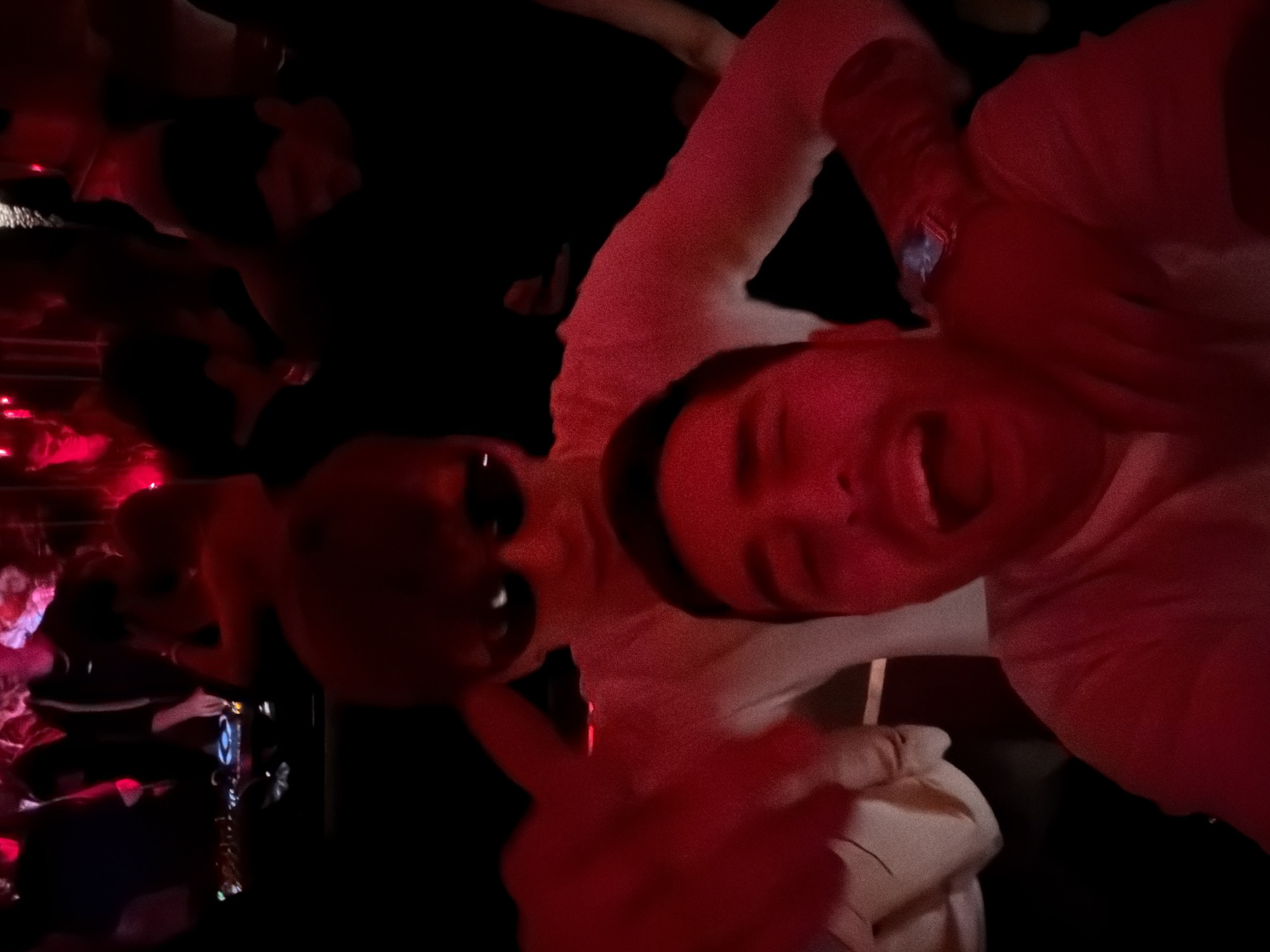 Two individuals pose for a selfie in a dimly lit environment, seemingly at a club or venue. The man on the right is smiling broadly with an open mouth. The person on the left is wearing sunglasses. The environment is dark with red lighting, and there are other people in the background, some with raised hands. In the background there appears to be a bar or counter. There is no visual information to identify the location as Moscow, Russia.
