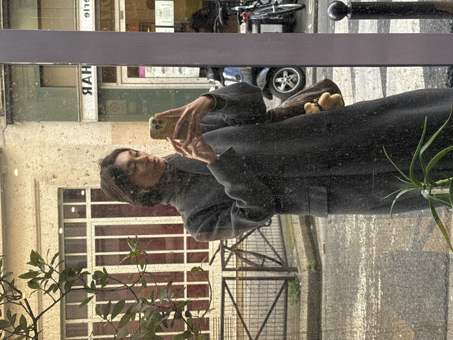 The image displays a woman's reflection capturing a selfie on an urban street in Paris, France. The woman, with dark hair and a dark grey turtleneck and long coat, holds a gold-colored smartphone directly towards her reflection. She wears gold hoop earrings, and a brown shoulder bag with a teddy bear charm is visible on her right side.
The reflected background reveals typical Parisian architecture: stone or stucco buildings. One prominent building exhibits a large window with vertical red bars. Adjacent to it, a shopfront displays reversed text, notably "ЯАН" (RAH) and "ЭІЯ" (EIE). On the street, a dark scooter with silver wheels is parked, alongside multiple bicycles. Black bollards are visible near the bottom right of the reflection. Green tree foliage extends into the upper left, partially obscuring a building. In the immediate foreground, at the base of the reflective surface, a potted plant with long, spiky green leaves is present. The reflective surface itself is visibly mottled with numerous small specks, consistent with dirt or water drops.