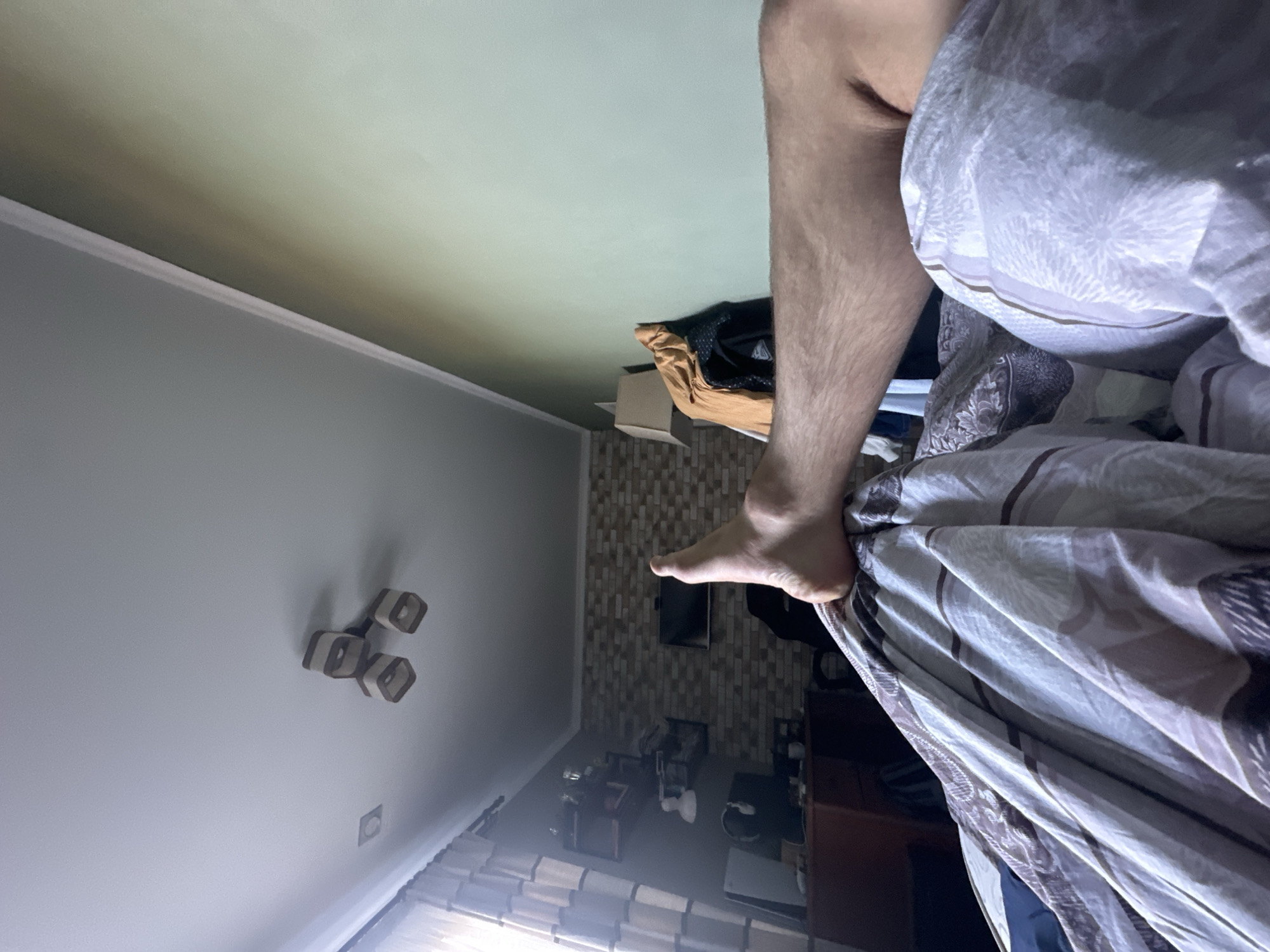 The image shows a bedroom interior. A leg and foot extend from under patterned bedding. In the background, a wall with brick-like tiling is visible, along with a television and various pieces of furniture including shelves and a desk. A light fixture is mounted on the ceiling, and a window with blinds is also visible. The location is unknown.