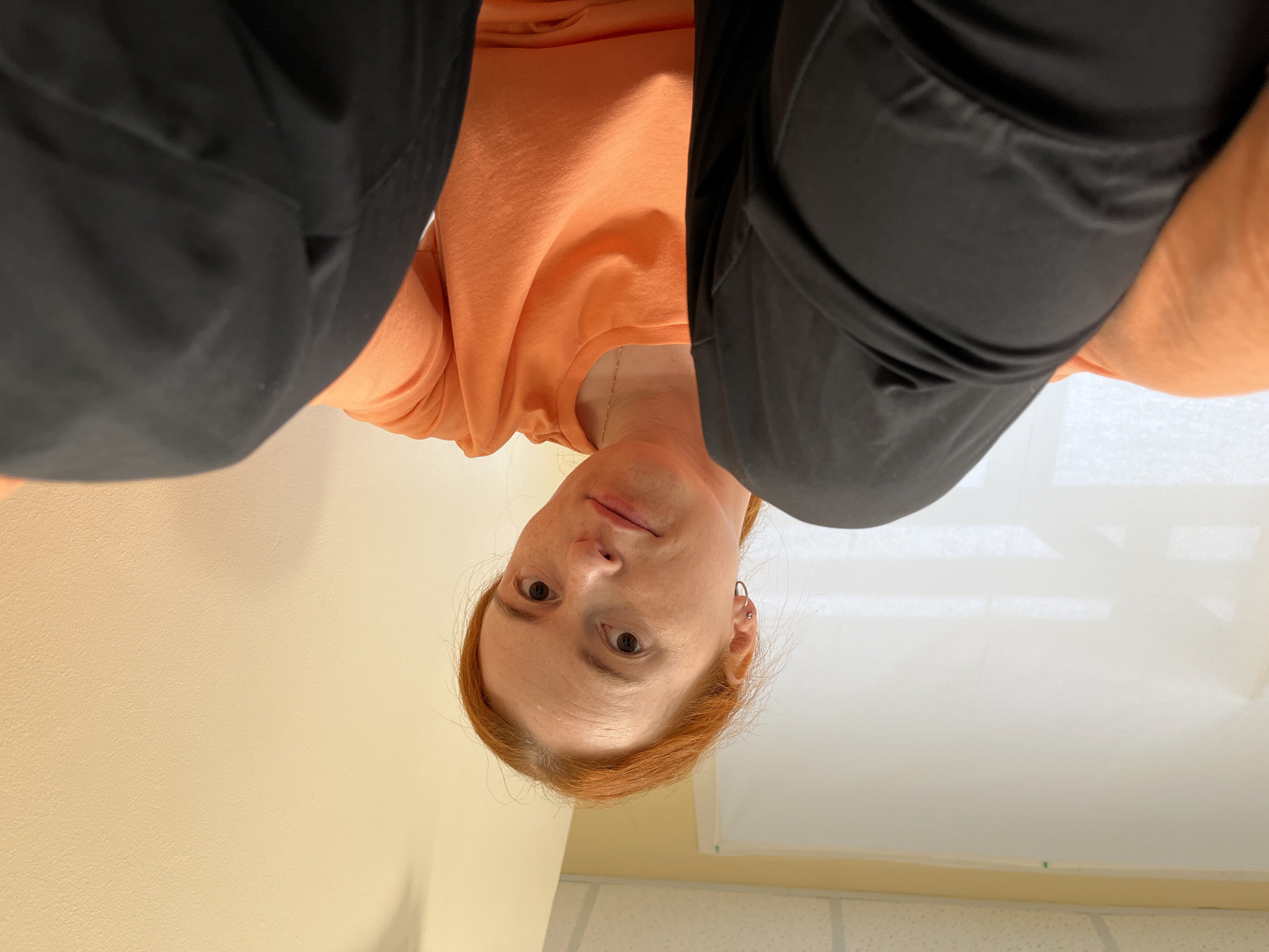 A young woman with red hair and freckles is sitting and looking at the camera. She is wearing an orange t-shirt and black pants. The background is a plain wall and a white screen. The image is taken from a low angle, looking up at the woman. The lighting is bright and natural, suggesting it is daytime. The location is not specified beyond Russia.