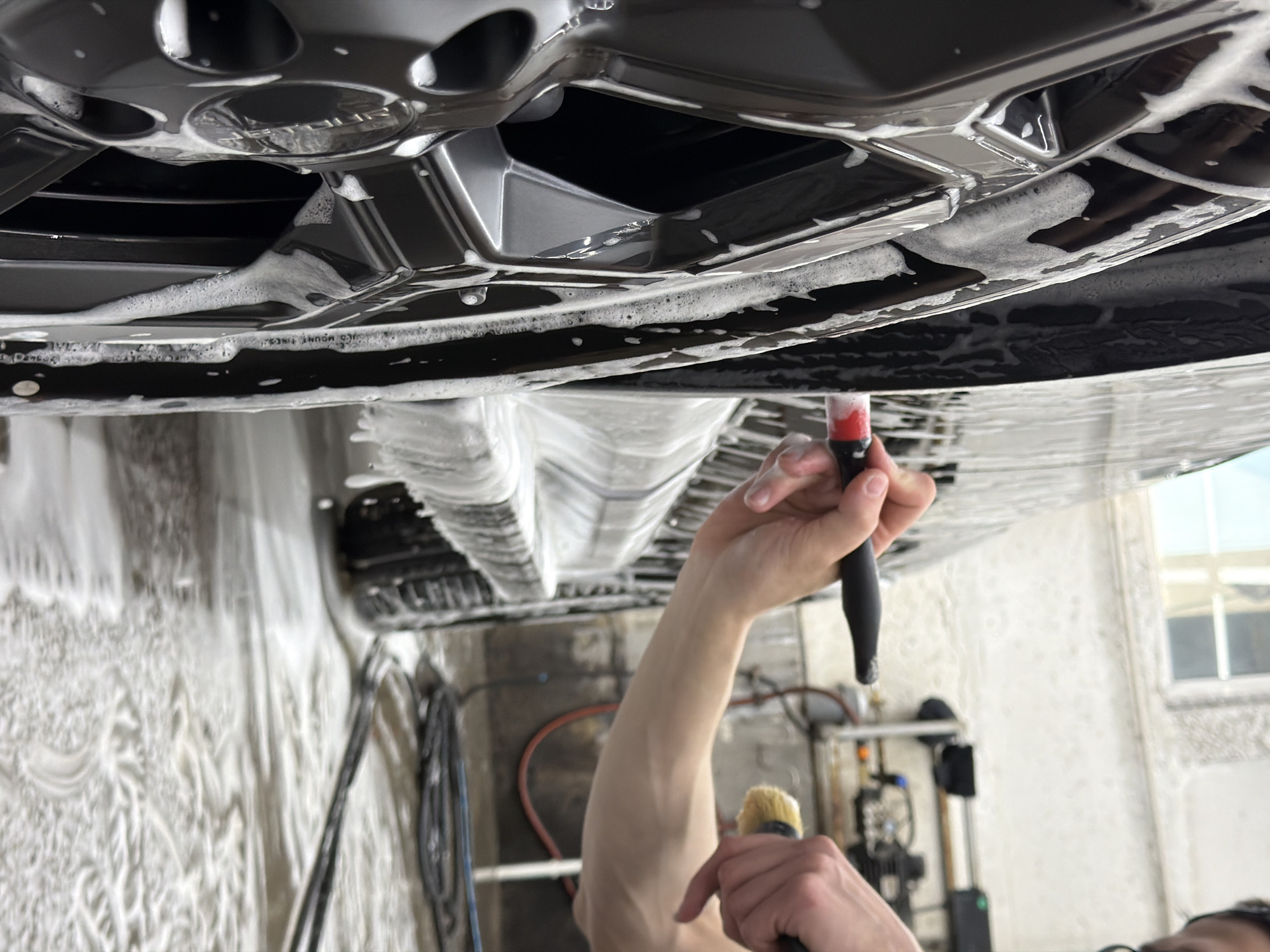 The image captures a car being washed, possibly at a car wash facility, as indicated by the soapy foam covering the vehicle and surrounding areas. The perspective is from below the car, looking upwards at the undercarriage. A person is visible, holding two brushes - one with a black handle and red tip, and another smaller one with yellow bristles. The car appears to have dark-colored wheels, and the environment includes industrial-looking equipment in the background, suggesting a professional setting. The image does not contain information to verify its location in the city of Krasnoyarsk, Russia.