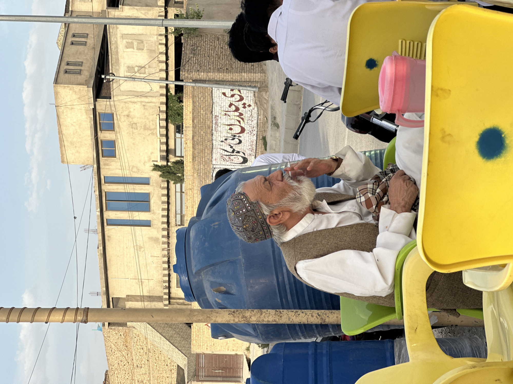 A medium-shot captures an elderly man with a white beard seated on a green plastic chair in what appears to be a public outdoor setting in Quetta City Tehsil, Pakistan. He is wearing a traditional patterned cap, a white shirt, and a brown vest. He holds a cigarette to his lips. He sits near blue plastic water containers.
Next to him is a yellow plastic chair with a pink plastic pitcher on it. Other individuals are partially visible, indicating a populated area. In the background, buildings and signage are visible. A sign written in Urdu advertises "The Children High School." The surrounding environment suggests an urban streetscape.