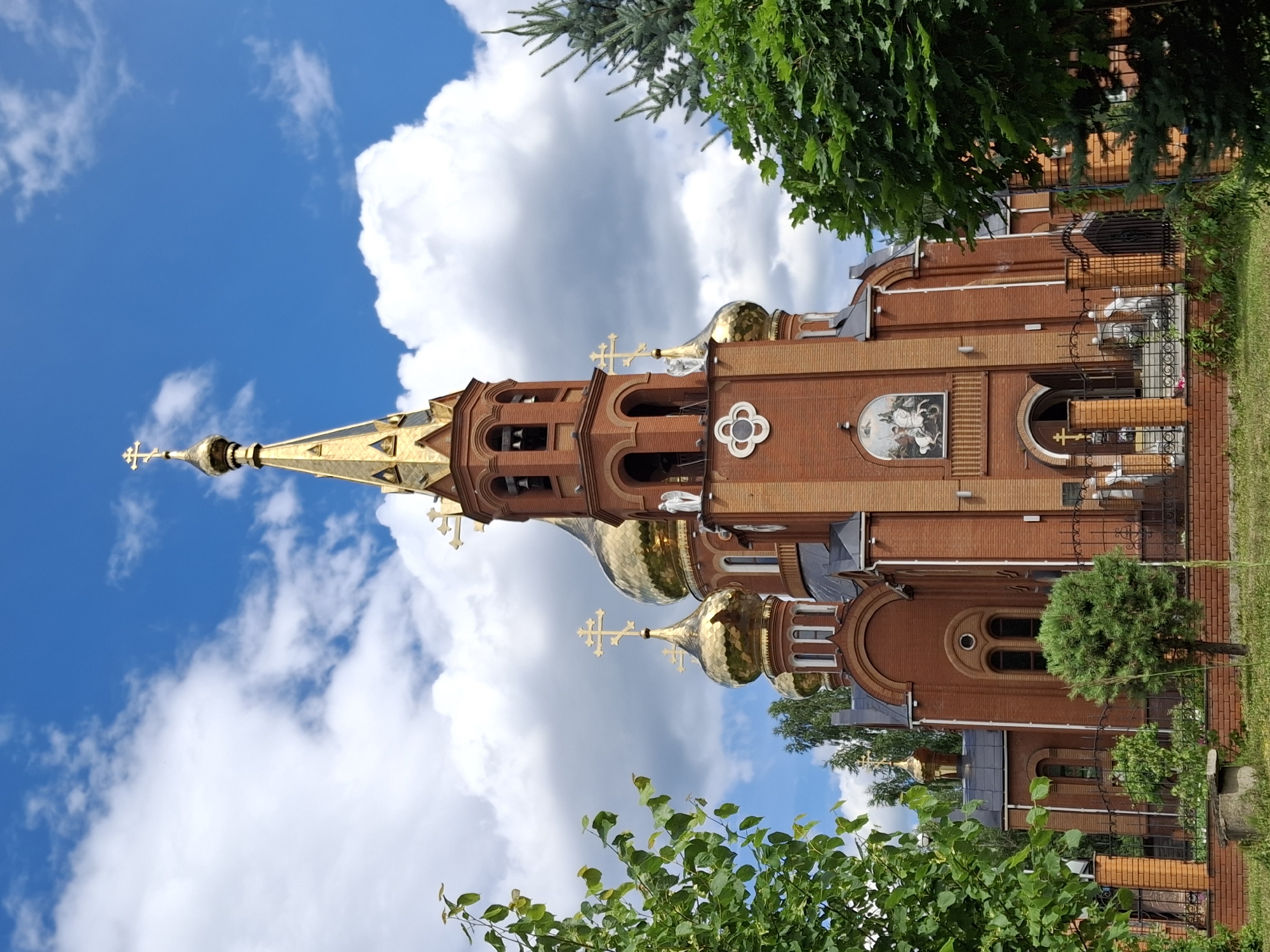 This is a vertical, eye-level, medium shot of an Eastern Orthodox church, likely in Chaikovsky, Russia. The church is constructed from red brick and features multiple golden domes topped with crosses. The main spire is tall and intricately designed, also adorned with a cross. Sunlight glints off the domes, indicating a bright day. The sky is a vibrant blue with fluffy white clouds. Lush green trees and foliage frame the church, suggesting it is a warm season, possibly summer. A small, well-maintained green bush is visible in front of the church. The church architecture includes arched windows and decorative elements, such as a circular floral motif on the brickwork and a mosaic depicting a religious scene above an arched doorway. A dark metal fence surrounds the property. The lighting and clear sky suggest it is daytime, likely mid-morning to afternoon. No people are visible in the frame.