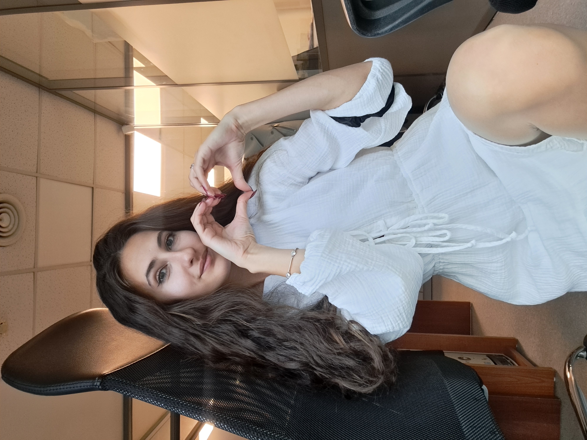 A young woman is posing indoors, making a heart shape with her hands near her face. She has long, wavy brown hair. She is wearing a white dress with long sleeves. A black office chair is visible behind her, and an office environment with windows and a ceiling with recessed lighting is in the background. There is no conclusive evidence to pinpoint the location to Sofia, Bulgaria.