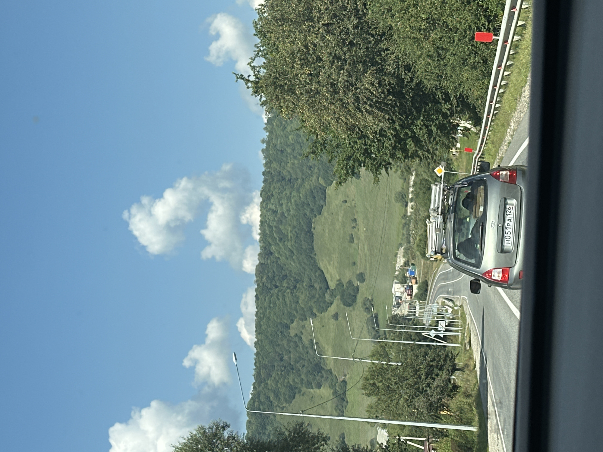 The image shows a view from inside a vehicle looking out at a road winding through a hilly, green landscape in the vicinity of Dausuz, Russia. A light gray car with a roof rack is visible ahead, driving on the road. The road is lined with streetlights and a metal guardrail. A lush green, tree-covered hillside forms the backdrop. The sky is blue with scattered white clouds. The scene appears to be a daytime drive through a rural or mountainous area.