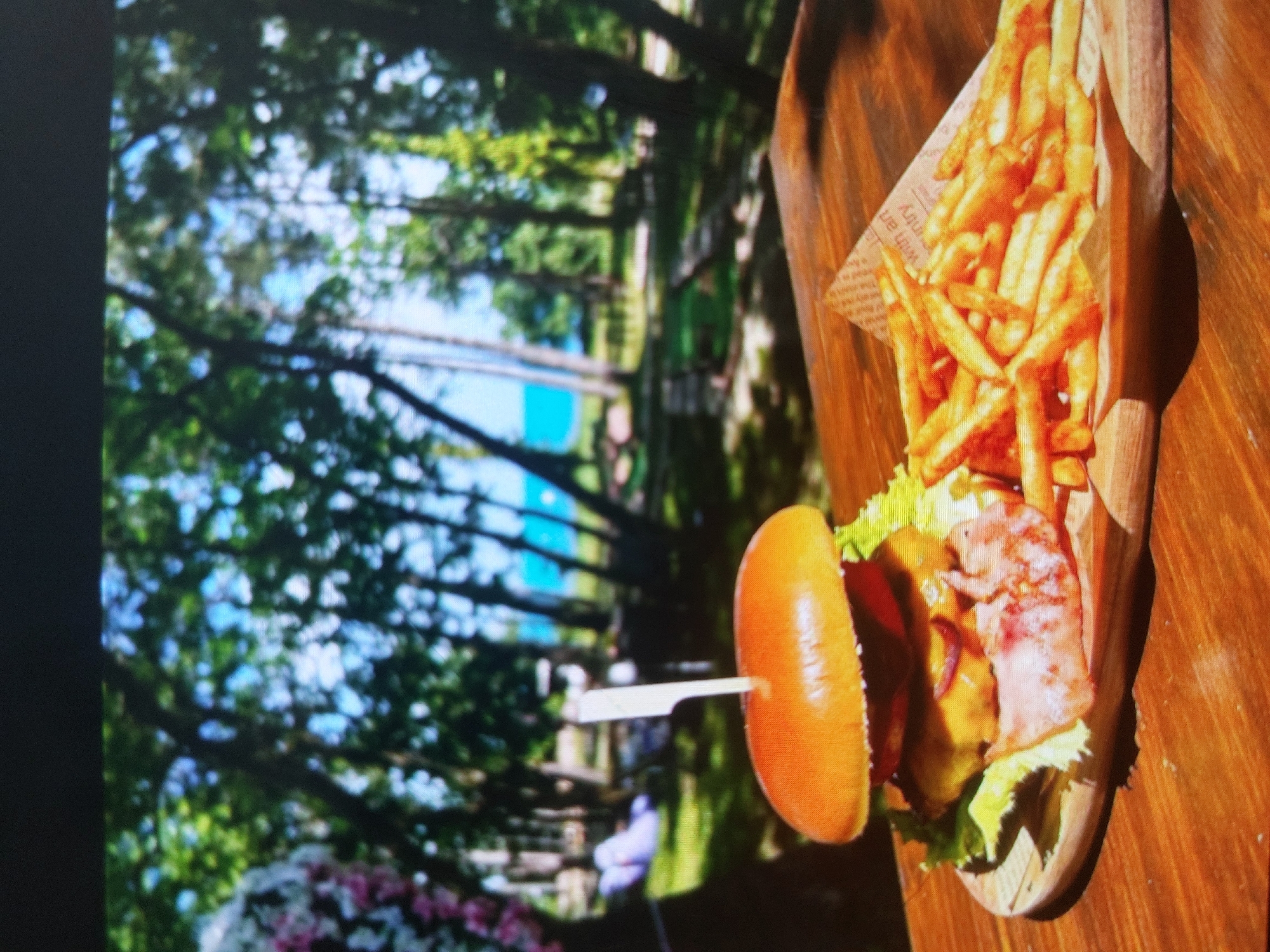 A partially visible burger and fries are served on a wooden platter. A skewer holds the top bun. The outdoor scene reveals green trees and a body of water. The burger is partially obstructed from view. The background appears to be outside and in Bulgaria. However, the location in Tsarevo, Bulgaria, is unverifiable.