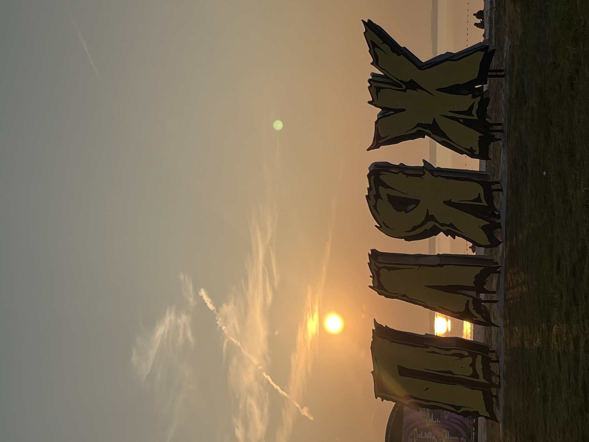 The image depicts a sunset over a body of water, likely a lake or sea, with the sky a warm gradient of orange and yellow. In the foreground, large, stylized yellow letters with dark outlines spell out "PLAYX". These letters appear to be three-dimensional installations. Behind the letters, the water stretches to the horizon, where a faint treeline is visible. The sun is a bright, glowing orb low in the sky, casting a reflection on the water. A few wispy clouds are present, one of which has a long, thin contrail. On the grass in front of the letters, there is a small, dark object, possibly a piece of litter. Further back, on the shoreline, silhouettes of people can be seen, some sitting and others standing. To the far left, a large, dark structure, possibly a stage or tent, is partially visible. The overall scene suggests an outdoor event or a public recreational area in Surakovo, Russia.