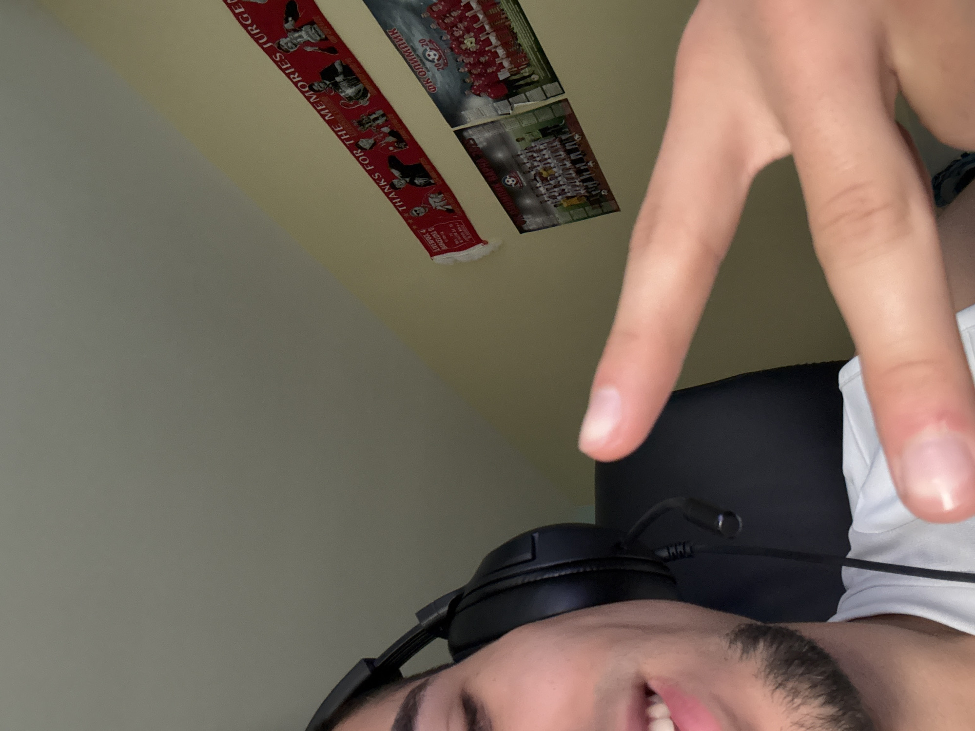 The image shows a man wearing a headset and giving a peace sign. In the background are items taped to a wall: a red and black Liverpool F.C. scarf with the words "THANKS FOR THE MEMORIES JURGEN" and images of FK Olympic Varna, a soccer team, indicating a possible location in Varna, Bulgaria. The man is wearing a white shirt. The environment appears to be indoors, possibly a bedroom.