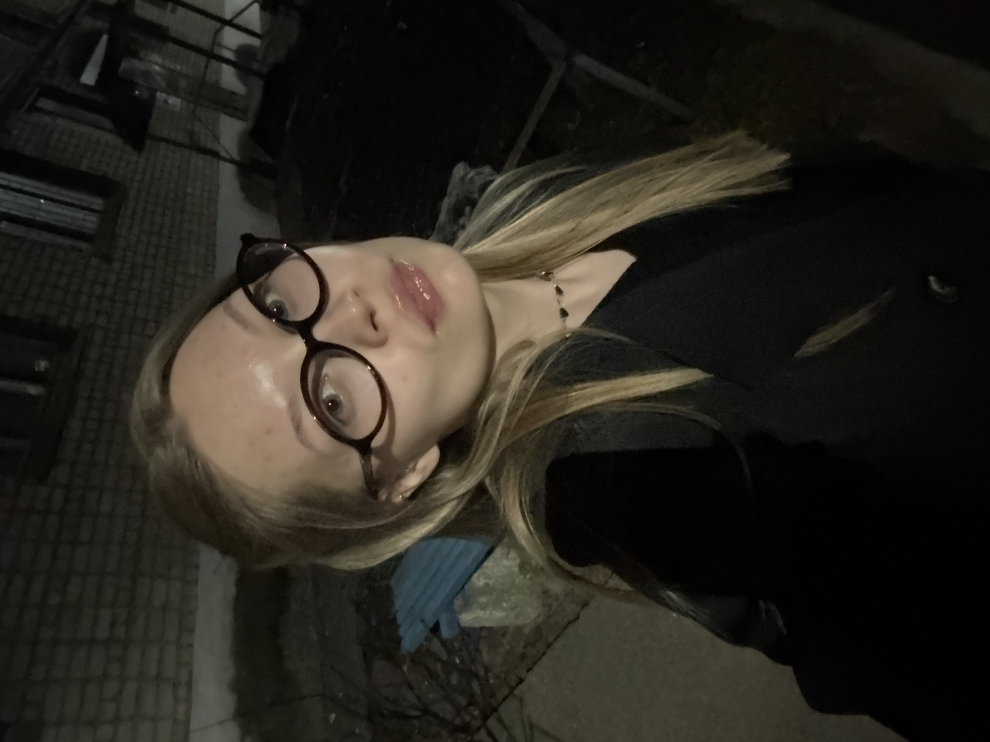 A vertical selfie shows a young individual, appearing to be female, with long blonde hair and dark-rimmed glasses, looking directly at the camera from an upward angle. The person is wearing a dark, possibly black, coat or jacket and a delicate necklace with small dark charms. Her lips are slightly parted and appear glossy. Her expression is neutral.
The image was taken at night, as indicated by the dim, dark surroundings, with the person's face being the primary illuminated area. The background features a brick-patterned surface, resembling either a building wall or pavement, along with dark, indistinct areas. In the lower part of the image, the ground shows more of the brick pattern, a faint white line, and a small, blurry blue object. The setting appears to be outdoors in an urban or suburban environment in Chaikovsky, Russia. There is no visible text.