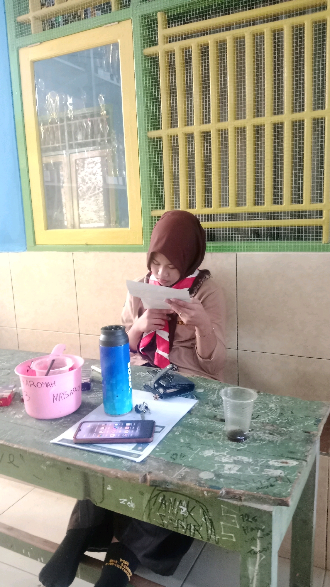 A young woman wearing a brown hijab and a school uniform, possibly a scout uniform, sits at a green wooden desk. She is holding a piece of paper and looking down at it. On the desk in front of her is a pink container with writing on it, a blue water bottle, a wallet, a smartphone, and a clear plastic cup with liquid. Behind her is a wall with beige tiles and a window with a yellow frame and green mesh. The scene appears to be indoors, possibly a classroom or common area in Waru, Indonesia. The desk has graffiti on it.