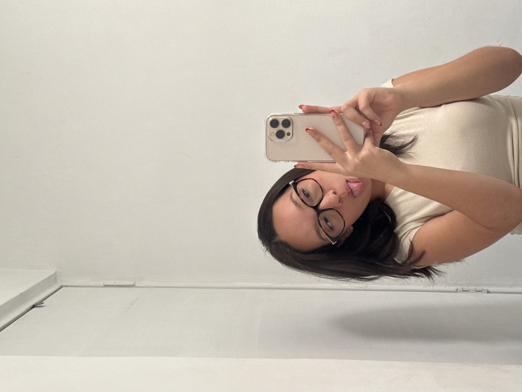 A young woman with dark hair and glasses is taking a selfie with a phone. She is wearing a light-colored top and has red nail polish. Her tongue is sticking out. The background is a white wall and a white door. The photo appears to be taken indoors. It is not possible to determine the location in Puebla, Mexico, from the image alone.