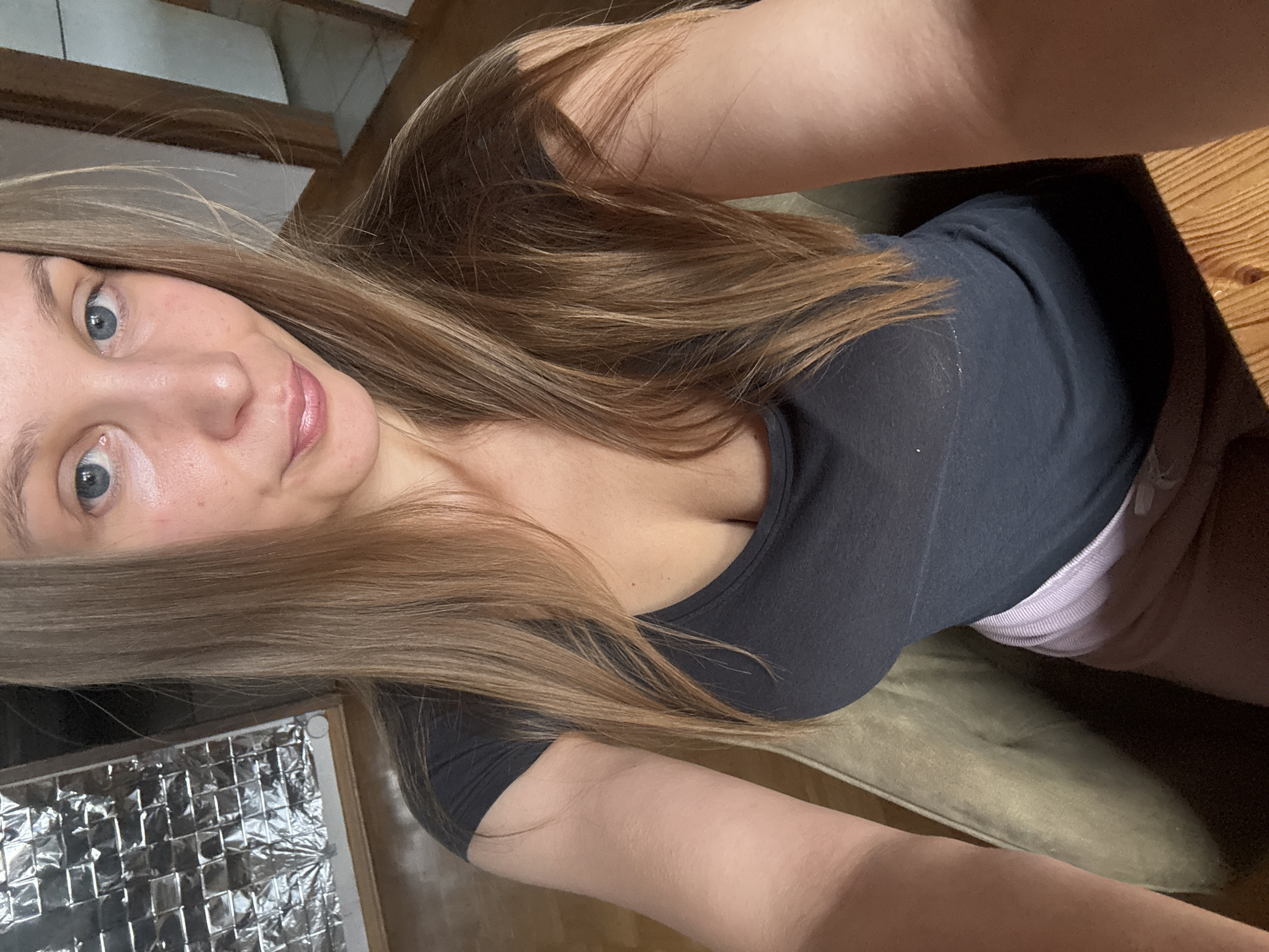 This is a first-person, vertical, medium shot of a young woman. She has long, light brown hair that falls over her shoulder and chest. Her face is turned towards the camera, with her blue eyes looking directly at the viewer. She has a slight smile on her lips, and her skin appears clear and lightly flushed. She is wearing a dark grey t-shirt that is somewhat sheer, revealing the pink and white striped band of her underwear underneath. She appears to be sitting, as her arm is resting on a surface and her legs are visible in the lower portion of the frame.
The background is blurred, but hints of a wooden table and a couch can be discerned. To the left of the frame, there is a reflective object that appears to be a heating element or insulation, possibly part of a heater. The lighting is somewhat dim, suggesting it might be evening or indoors. There is no visible text in the image. The overall mood is casual and intimate.