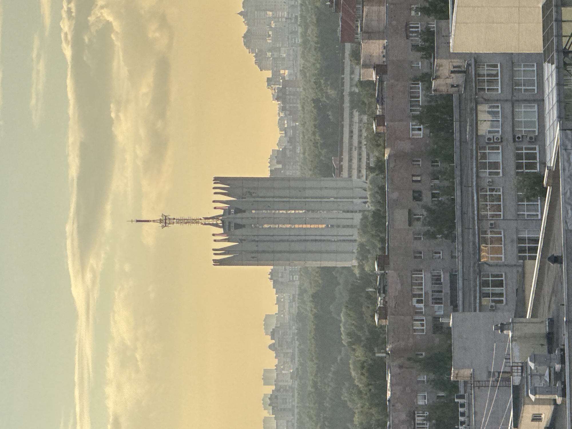 This is a medium-shot, vertical photograph of a cityscape taken from an elevated vantage point, likely at sunset given the warm, hazy lighting and the yellowish sky with scattered clouds. The most prominent structure in the mid-ground is a tall, Brutalist-style building with a distinctive crown-like top, featuring pointed vertical elements. A radio tower with antennae extends from the very top of this building.
In the foreground, there are several multi-story residential buildings with flat roofs. These buildings are constructed of brick and concrete, with numerous windows. Some roofs have visible ventilation shafts, chimneys, and other utility structures.
Behind the main Brutalist building, the cityscape stretches into the distance, showing a dense collection of urban structures, including apartment blocks and other buildings, all softened by atmospheric haze. A dense line of trees separates the foreground residential buildings from the more distant urban landscape. The overall impression is one of a sprawling urban environment under a warm, diffused light. There are no people or discernible actions visible in the scene.