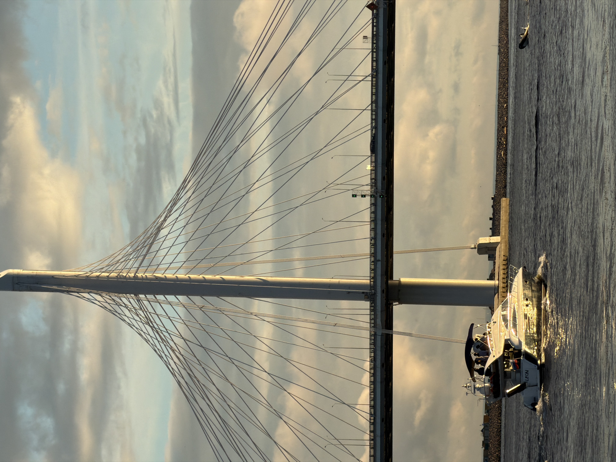 The image depicts the Western High-Speed Diameter (WHSD) bridge in Saint Petersburg, Russia, during what appears to be sunset. The massive cable-stayed bridge is a dominant feature, with its tall, slender white pylon and radiating cables against a cloudy sky tinged with orange and yellow hues. Below the main bridge deck, a modern, white motor yacht with several people onboard is cruising on the water, leaving a wake. The yacht has a dark canopy over the main seating area and a Russian flag displayed at the stern. In the distance, on the right side of the frame, a smaller, dark inflatable boat is visible. The water is choppy, reflecting the warm light from the setting sun. The background shows a stone embankment and structures in the distance, suggesting an urban or semi-urban waterfront. The overall scene is one of architectural grandeur combined with leisure activity on the water, captured in the soft, warm light of late day.