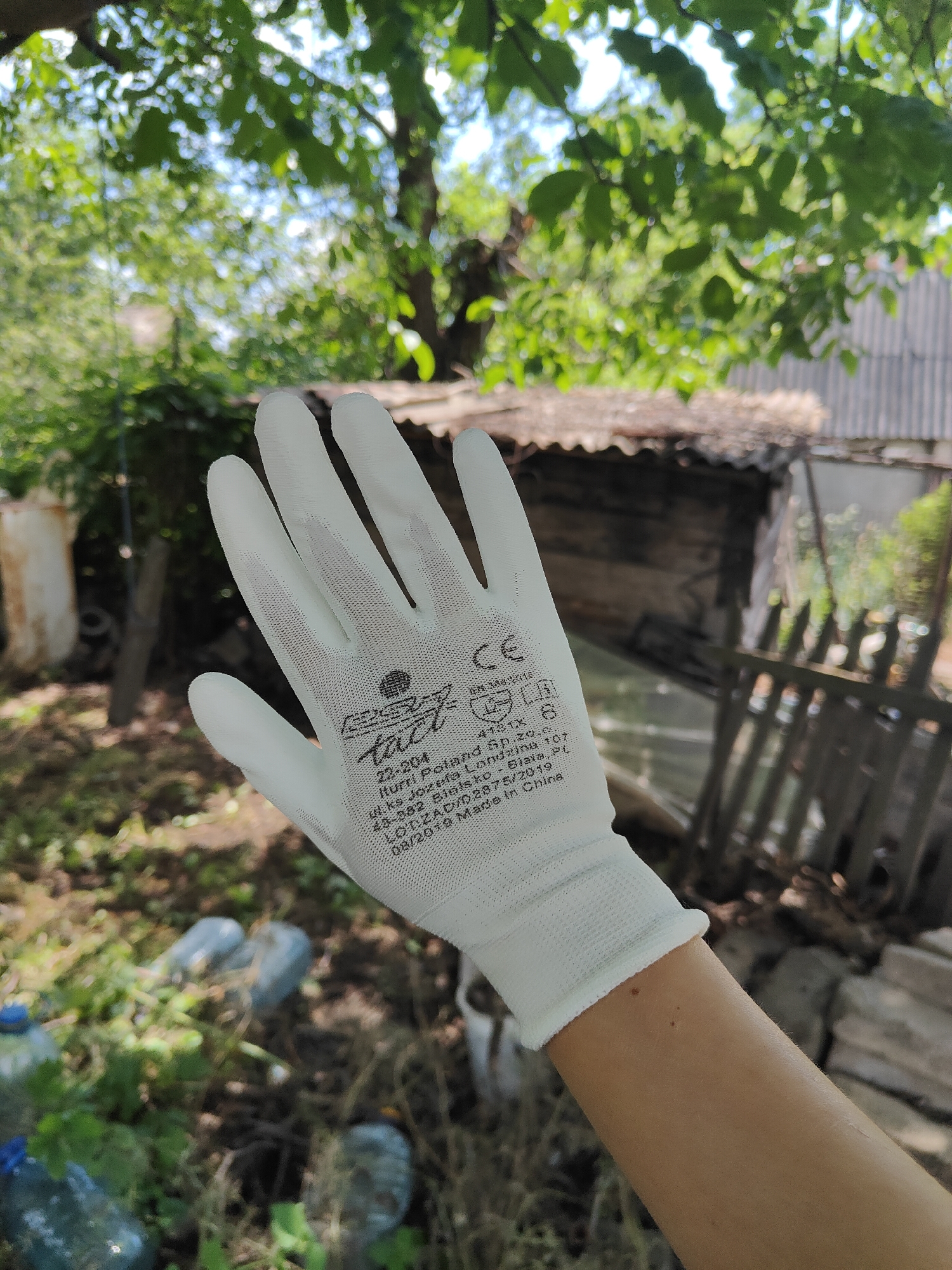 A person's left hand, wearing a white glove, is shown in the foreground, held up in front of a blurred background. The glove has black lettering and symbols printed on it. The background depicts an outdoor scene with trees, foliage, a rustic structure, and some scattered plastic bottles on the ground. It is daytime and the lighting suggests a sunny environment. The scene appears to be in a rural or semi-rural area, possibly a backyard or garden. There are no discernible landmarks that specifically place it within Kryvyi Rih, Ukraine.