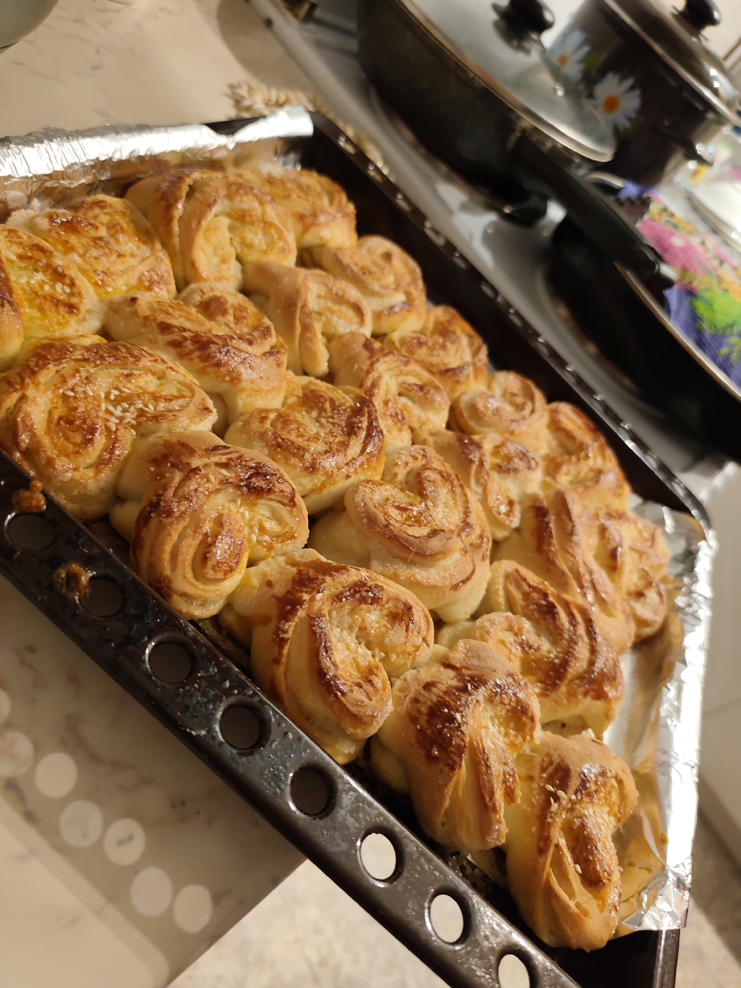 The image displays a dark metal baking tray filled with approximately two dozen golden-brown, spiral-shaped pastries or buns. The tray is partially lined with crumpled aluminum foil along its left edge. The baked items exhibit a shiny glaze, and many are topped with visible white sesame seeds or granulated sugar crystals. The pastries are arranged closely together on the tray, occupying the majority of the frame. In the blurred background, a light-colored countertop is visible, alongside two dark-colored cooking pots with lids, one of which features a white daisy floral pattern. A multi-colored textile, likely a kitchen towel, displaying blue, pink, and green hues, is partially visible near the pots. No individuals are present. The scene is an indoor domestic setting, specifically a kitchen. The exact location within the city of Chaikovsky, Russia, cannot be determined from the visual evidence.