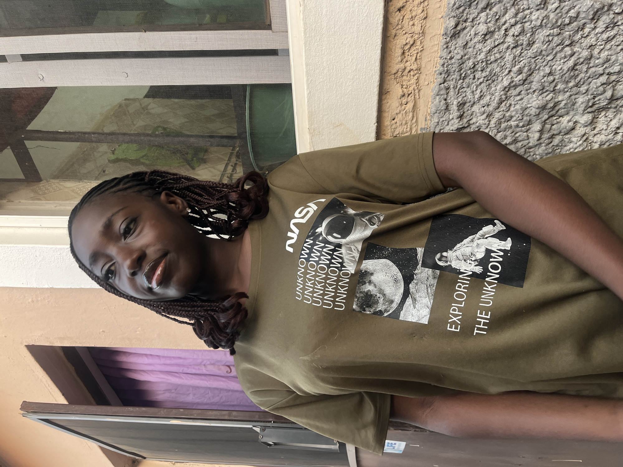 A young Black woman is pictured wearing a light brown t-shirt with a NASA-themed design featuring images of space exploration and the words "UNKNOWN" and "EXPLORING THE UNKNOWN". The t-shirt has a graphic print including an astronaut and a moon. Her hair is styled in brown braids with white and black beads or accents near her face. She is looking at the camera with a slight smile.
The background is a mix of interior and exterior elements. Behind her, there is a window with a screen, suggesting an indoor setting, along with parts of a wall that appear to be outdoors. There is a hint of a purple curtain visible near the bottom left of the frame. The location is Abuja, Nigeria, based on the available information.