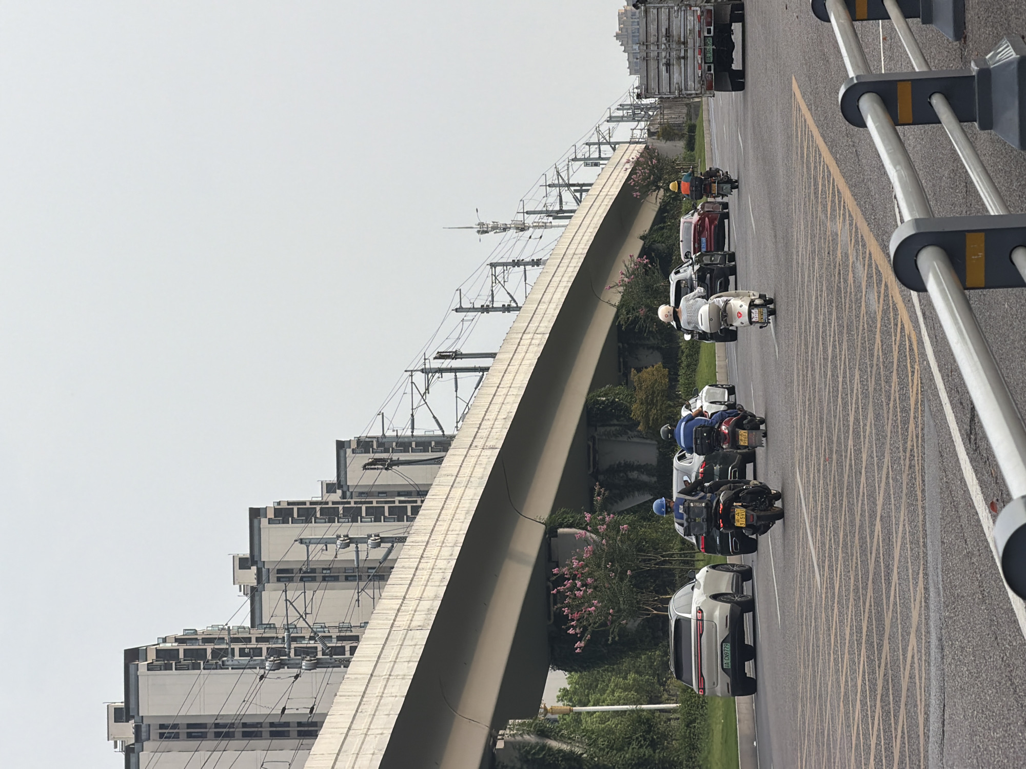 The image shows a multi-lane road in the city of Jiangjin, China, with a highway overpass above. Several vehicles are visible on the road, including cars, motorcycles, and a truck. On the left side of the road, there are tall buildings and what appears to be a train track or elevated highway with overhead power lines. The sky is overcast.
More specifically, there are four motorcycles and three cars in motion on the road. Two of the motorcycles are in the foreground, ridden by individuals wearing helmets and what appear to be delivery bags. A white SUV is in the left lane, followed by a dark-colored car. Further ahead, another motorcycle with a rider in an orange helmet is visible, along with a red car. In the right lane, a large truck is traveling away from the viewer.
The overpass is supported by concrete pillars, and vegetation is growing on some of them. The buildings in the background are modern, multi-story structures, characteristic of urban development. The road is marked with yellow and white lines. The overall scene depicts a typical urban transportation environment in Jiangjin.