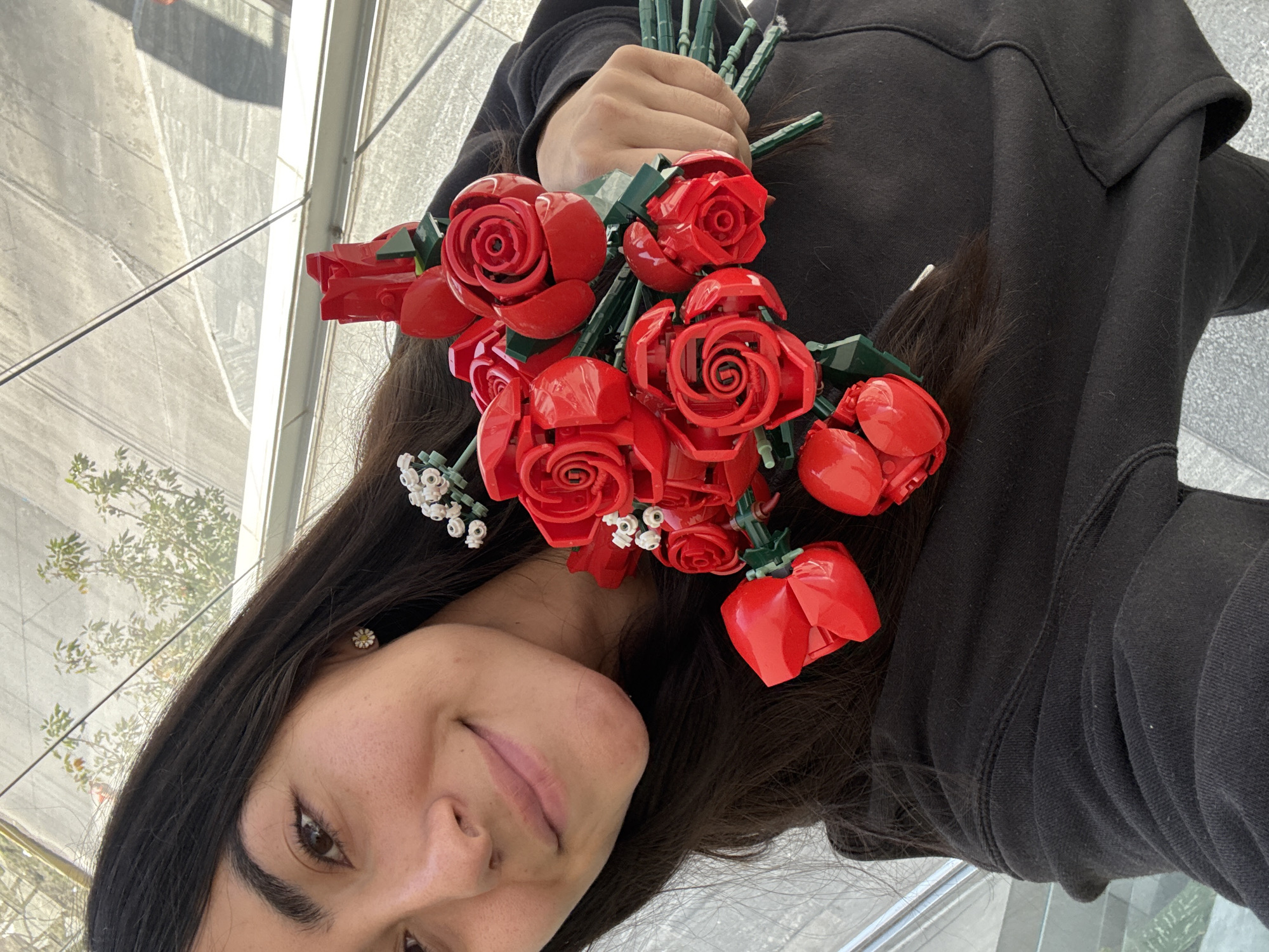 A young woman is captured in a selfie-style photograph, holding a vibrant bouquet of red, block-built roses, likely from a Lego botanical set. Her dark hair cascades around her face and over her shoulders. She wears a black long-sleeved top, possibly a hoodie or sweatshirt, and has a small, daisy-like stud earring in her left ear. A gentle, subtle smile plays on her lips, suggesting contentment.
The bouquet of artificial roses features intricate red petals, green stems, and small white accent flowers, all constructed from building blocks. Her left hand is visible, gripping the green stems of the bouquet.
The background, visible behind and above her, indicates an outdoor setting, likely in an urban or semi-urban area of Puebla, Mexico. Reflections in large glass panels suggest a modern building, showing glimpses of a grey paved surface and green foliage from trees or bushes. The bright lighting suggests it is daytime, possibly a sunny morning or afternoon. The overall mood of the image is positive and cheerful. There is no visible text in the image.