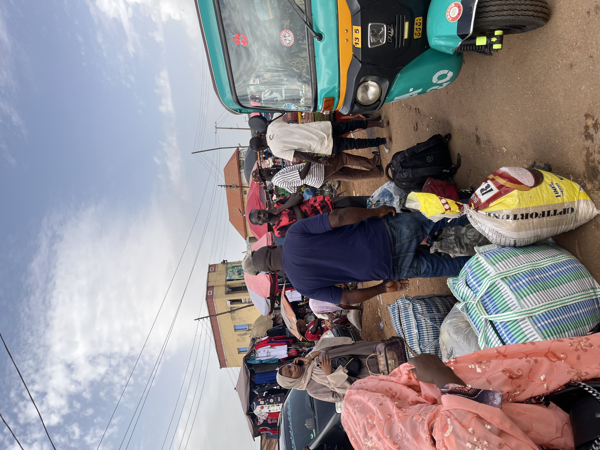 The image depicts a bustling market scene in Rafin Par, Nigeria. In the foreground, several people are gathered amidst a collection of large bags, indicating travel or commerce. A woman in a light brown hijab and a patterned pink garment is talking on her phone, standing near a parked car. To her left, a man in a dark blue t-shirt and jeans stands with his back to the camera, also surrounded by bags.
On the right side of the image, a bright teal and yellow auto-rickshaw, a common mode of transport in the region, is prominently displayed. The rickshaw has various stickers and a license plate with the number "13 5". Behind the rickshaw, more individuals are interacting, some appearing to be vendors or customers at market stalls.
In the background, colorful market stalls are set up with clothing and other goods hanging for display, covered by awnings and umbrellas. Buildings with yellow and red facades are visible, suggesting a mix of commercial and residential structures. The sky above is partly cloudy, with visible power lines crisscrossing the frame, characteristic of an urban or semi-urban environment in Nigeria. The ground is dusty and appears to be a dirt road or an unpaved market area. The overall atmosphere suggests a lively and active marketplace.