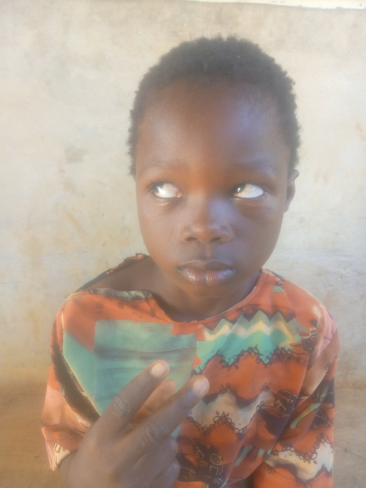 A young, dark-skinned child with short, dark hair is shown from the shoulders up against a light tan wall. The child is wearing a brightly colored shirt with orange, teal, and black patterns. The child is looking to the upper right, with a slight smirk. The child's left hand is raised, displaying the index and middle fingers in a "peace sign" gesture.
Location: Unknown, Unknown.