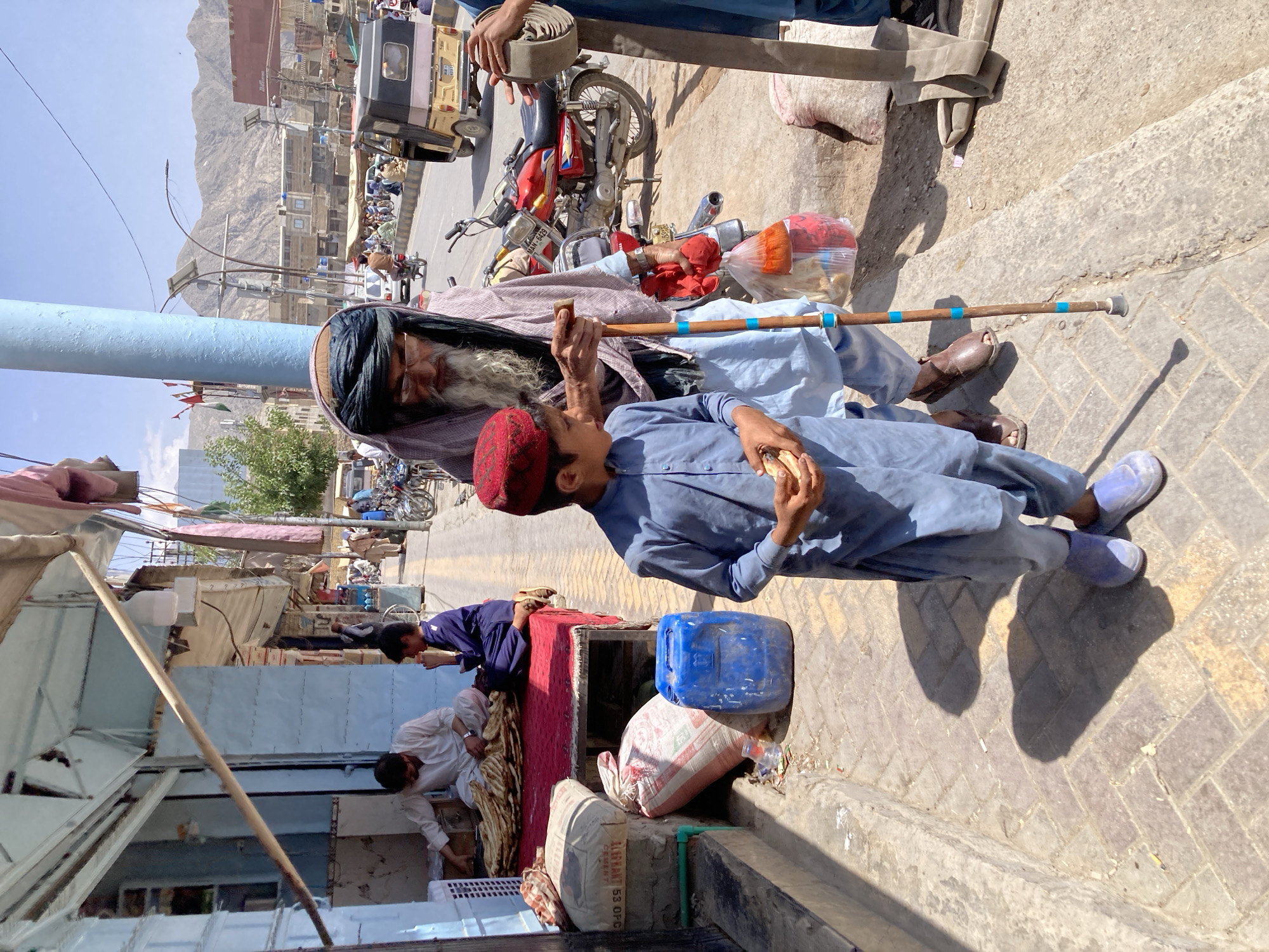 An old man with a long white beard wearing a black turban and light blue clothes sits on the ground with a cane. A young boy in a red cap and light blue clothes stands next to him eating bread. They are on a paved sidewalk next to a street with motorcycles, cars, and people. There are buildings and mountains in the background. In the foreground, there are blue and white containers, bags, and people under a canopy. The location is Quetta City Tehsil, Pakistan.