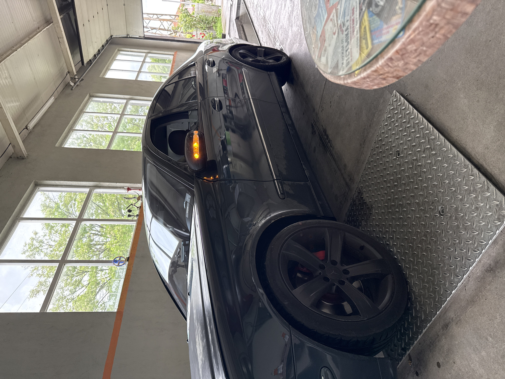 A black Mercedes sedan is parked on a diamond-plate service lift inside a garage, likely for maintenance or repair. The car's left turn signal is illuminated. The garage interior is lit by natural light filtering through a series of multi-pane windows along the left wall, which shows green trees in the background. The location is Stara Zagora, Bulgaria. A round table with a patterned top partially obstructs the view in the upper right corner. The walls of the garage are painted in neutral tones.