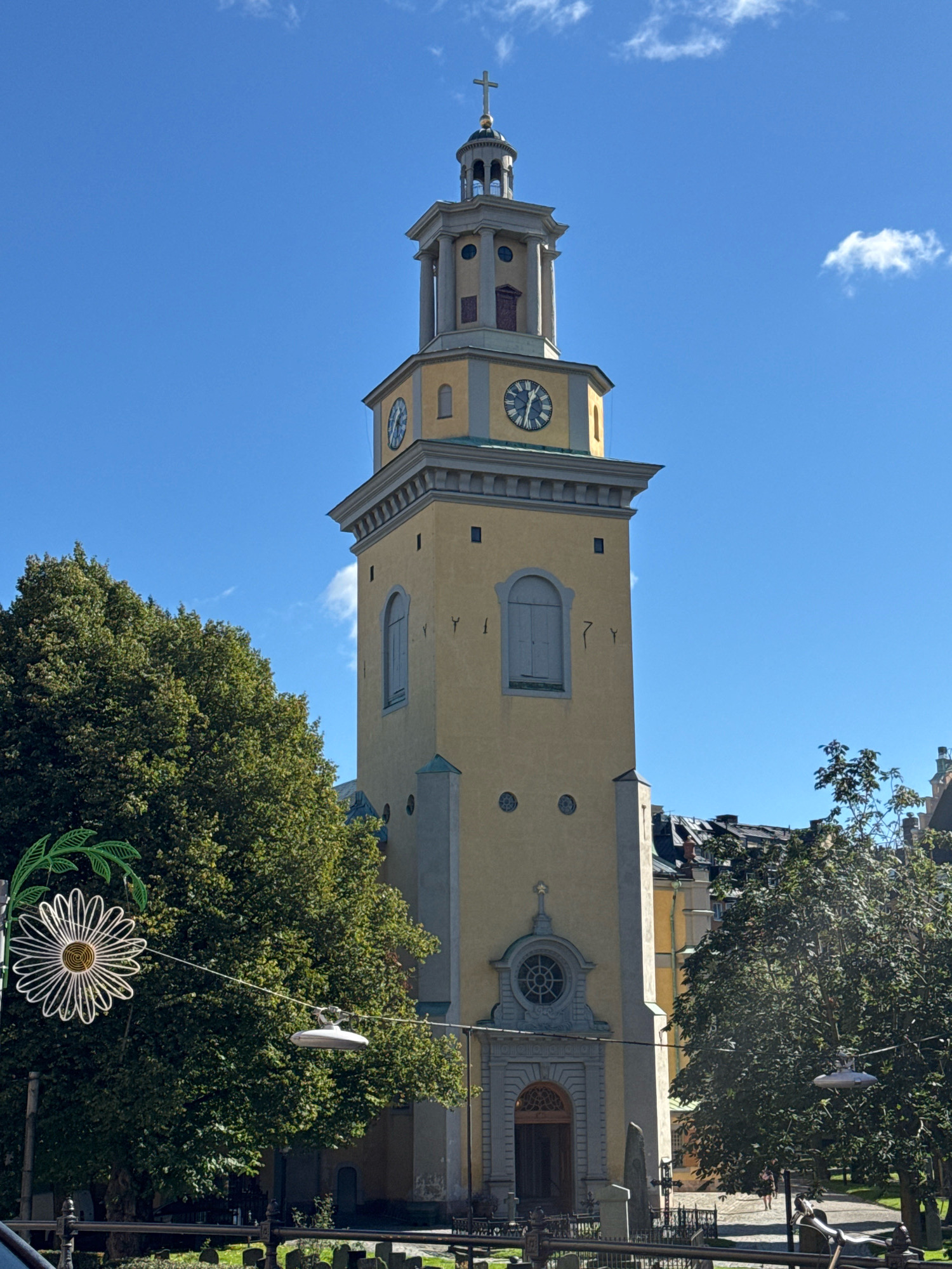 The image captures a close-up, low-angle shot of the yellow bell tower of a church, likely St. James's Church (Sankt Jacobs kyrka) in Stockholm, Sweden, given its distinctive architecture and the presence of similar surrounding buildings. The tower is tall and rectangular, with a grey cornice and a dome-like top crowned with a cross. It features several clocks on its upper section, Roman numerals and hands visible on at least one. The tower is adorned with arched windows and smaller circular openings. Below the tower, the entrance to the church is visible, featuring a large, ornate wooden door beneath a circular window with a geometric pattern.
To the left of the tower, a large, lush green tree dominates the frame, its branches heavy with leaves. A decorative, large white flower-shaped light fixture, resembling a daisy, hangs on a pole to the left of the tree. A string of lights, with a single white fixture visible, stretches horizontally across the image in front of the tree.
In the background, to the right of the church tower, more buildings with pitched roofs and dormer windows can be seen, suggesting an urban environment. The sky is a clear, vibrant blue with a few scattered white clouds. A portion of a graveyard with gravestones and fencing is visible in the foreground, partially obscured by the lush greenery of the tree and decorative elements. The overall scene suggests a bright, sunny day in a historic urban setting.