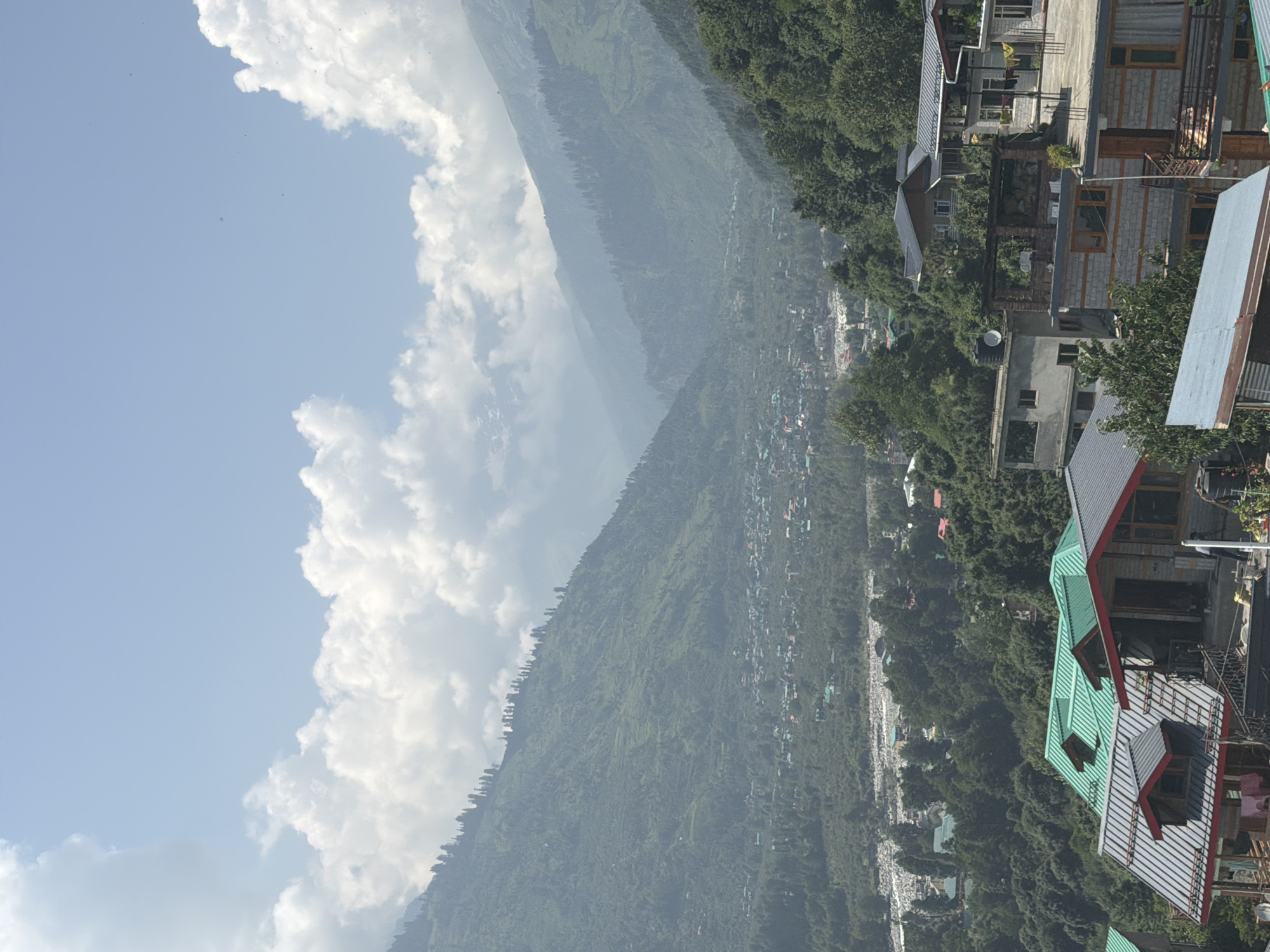 The image is a wide shot taken from a high vantage point, looking down into a valley that is part of Vashisht, India. The sky is a pale blue with scattered, fluffy white clouds. In the foreground, there are several multi-story buildings with pitched roofs, some covered in green metal and others in grey metal. These buildings appear to be homes or guesthouses, with visible windows, balconies, and what look like laundry hanging to dry. One building has a red trim around some of its windows and a darker grey roof.
Beyond the immediate foreground buildings, the landscape descends into a densely wooded valley. Numerous trees of varying shades of green cover the slopes. A town or village is nestled within the valley, characterized by a collection of smaller buildings with some having red or green roofs. A river or stream, appearing rocky and wide, runs through the valley floor.
The background of the image is dominated by large, verdant mountains that rise steeply from the valley. The mountain slopes are covered in dense vegetation and trees, with some appearing terraced. The haze in the distance suggests a significant depth to the valley and the mountainous terrain. The overall impression is a serene, rural landscape with human habitation integrated into the natural environment.