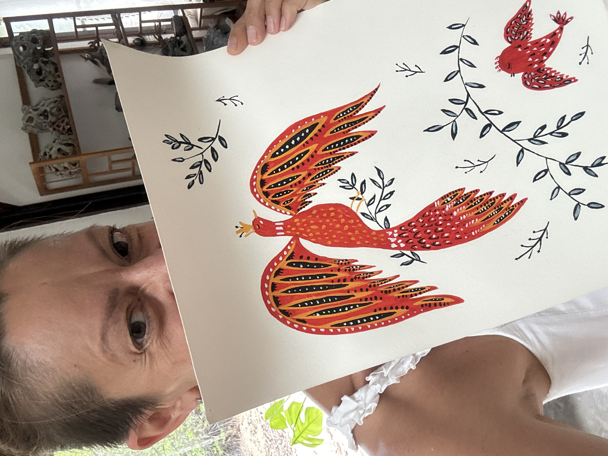 The image captures a person holding up a piece of artwork, with the photograph itself rotated 90 degrees clockwise. A woman's face is partially visible on the left side, showing her eyes, forehead, and ear. She has short, dark hair and fair skin, and is wearing a white top with ruffled straps.
The artwork she is presenting is a vibrant painting on a light-colored surface, likely paper or canvas. It features two stylized birds, predominantly red and orange, with intricate black and yellow patterns on their wings and bodies. The central, larger bird has elaborate, spread wings and a small golden crown on its head, facing left. A smaller, simpler red bird with white accents is depicted in the upper right, appearing to fly downwards. Scattered around the birds are decorative sprigs of black or dark gray leafy branches.
The background reveals an indoor setting with ample natural light. To the left of the person's head, a wooden structure resembling a shelf or room divider is visible, displaying several irregular, dark, rocky or coral-like decorative objects. To the lower right, out-of-focus green foliage suggests a window or open area looking out onto a natural, possibly tropical, environment, consistent with the specified location of Ko Pha Ngan, Thailand. The bright lighting indicates that the photo was taken during the day. No text is visible in the image.