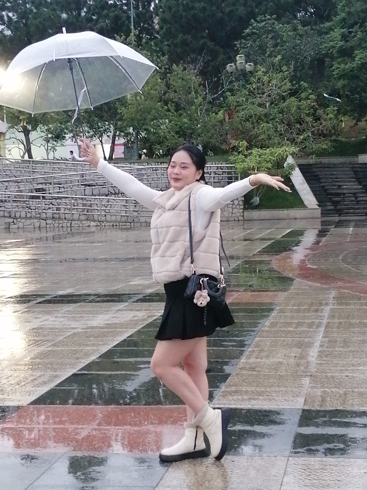 In Da Lat, Vietnam, a woman stands on a wet, tiled outdoor plaza, holding a transparent umbrella. She is wearing a white long-sleeved shirt, a beige faux fur vest, a black mini skirt, and white ankle boots. Her arms are outstretched, and she is looking to her right. A small black purse with a teddy bear charm hangs from her shoulder. The plaza appears to be a public space with stone stairs and railings visible in the background. Trees and foliage are also present, suggesting an outdoor setting in a city park or public square. The wet surface of the plaza indicates recent rain.