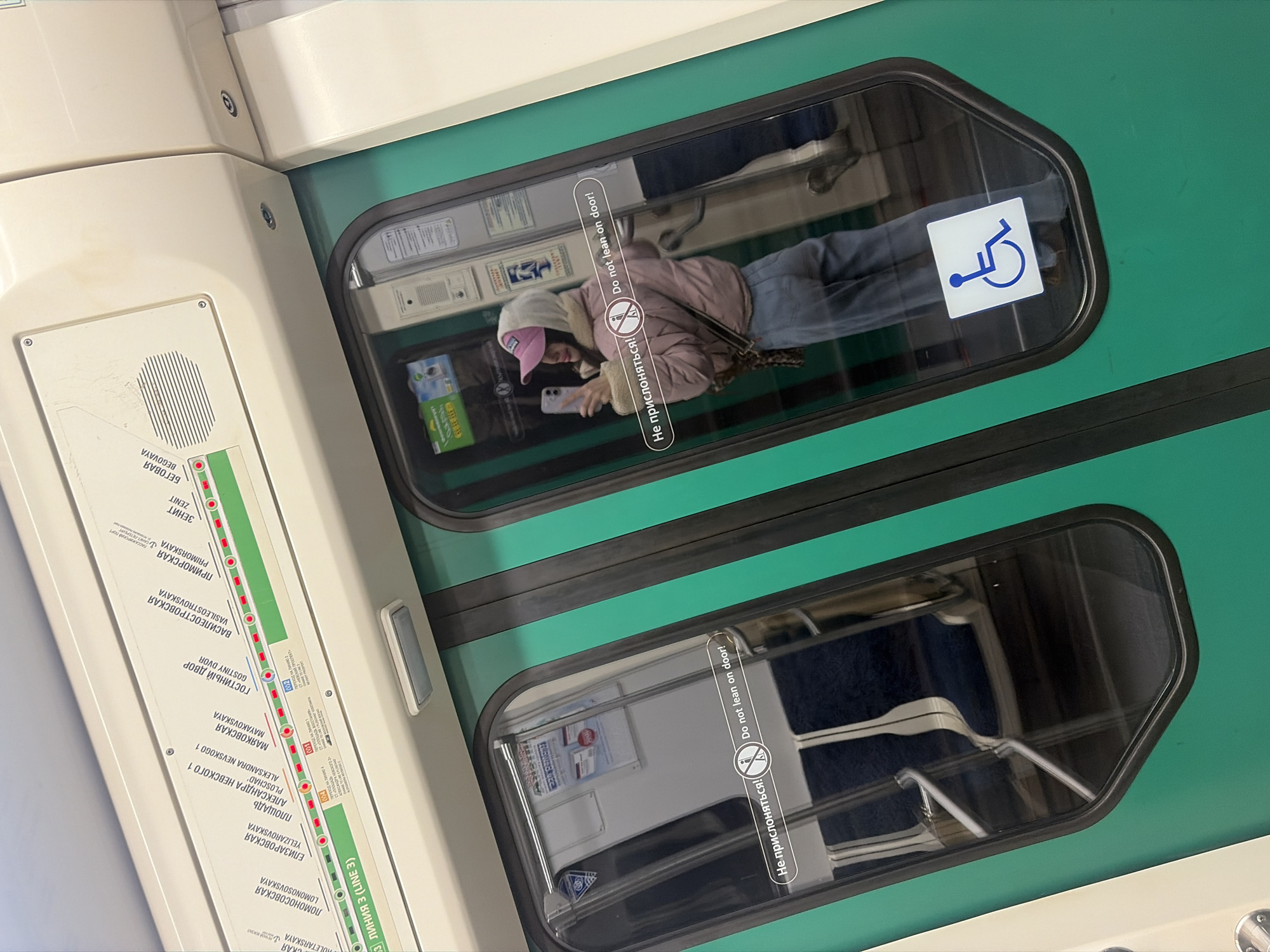 The image depicts the interior of a subway car in Saint Petersburg, Russia. It's taken from an angle, showing a segment of the car's wall with a train line map and two windows.
The train line map indicates the subway line 3, with stations listed in Russian and English, including "Lomonosovskaya", "Yelizarovskaya", "Ploschad Alexandra Nevskogo 1", "Mayakovskaya", "Gostiny Dvor", "Vasileostrovskaya", "Primorskaya", and "Zenit".
The upper window reflects the interior of the subway car and a passenger. A young woman wearing a pink hat and a pink jacket is looking at her smartphone. A sticker with a wheelchair symbol is on the upper-right corner of the window.
The lower window provides a view of the seating inside the car, revealing blue-colored chairs. Stickers in both windows read "Do not lean on the door!" in both Russian and English.
The environment appears clean and functional, typical of public transportation systems.