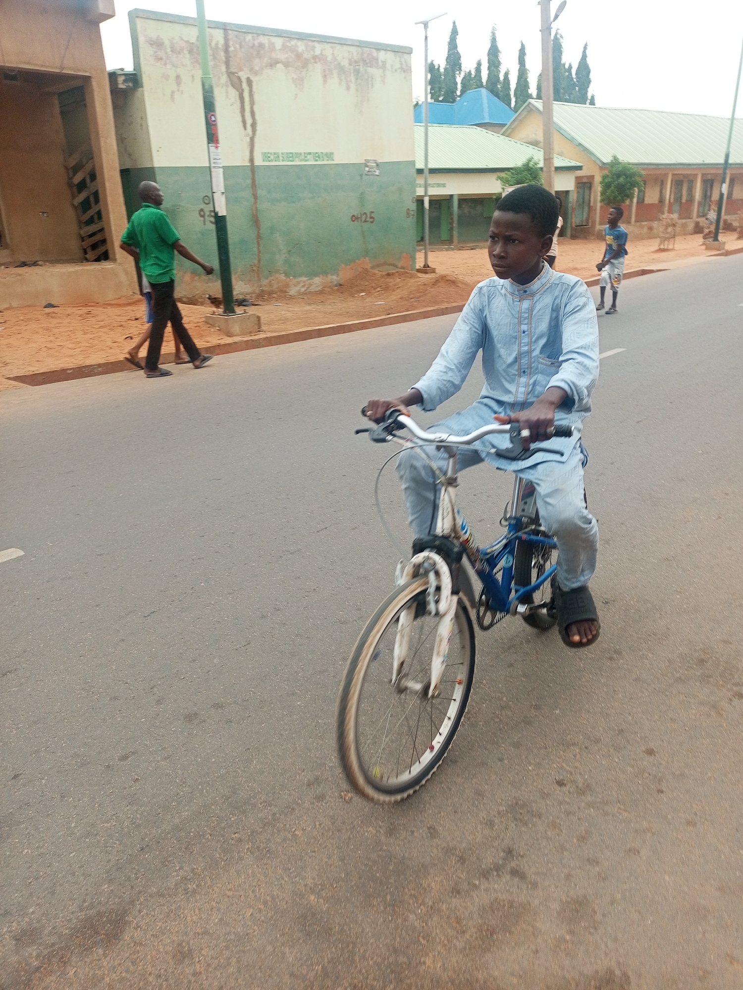 A young person wearing a light blue outfit and dark sandals is riding a bicycle on an asphalt road. Two individuals walk on the shoulder of the road to the left of the cyclist. A large building with green accents and faded writing along the side sits parallel to the road. The architecture and the environment suggest that this is a scene in Gombe, Nigeria.