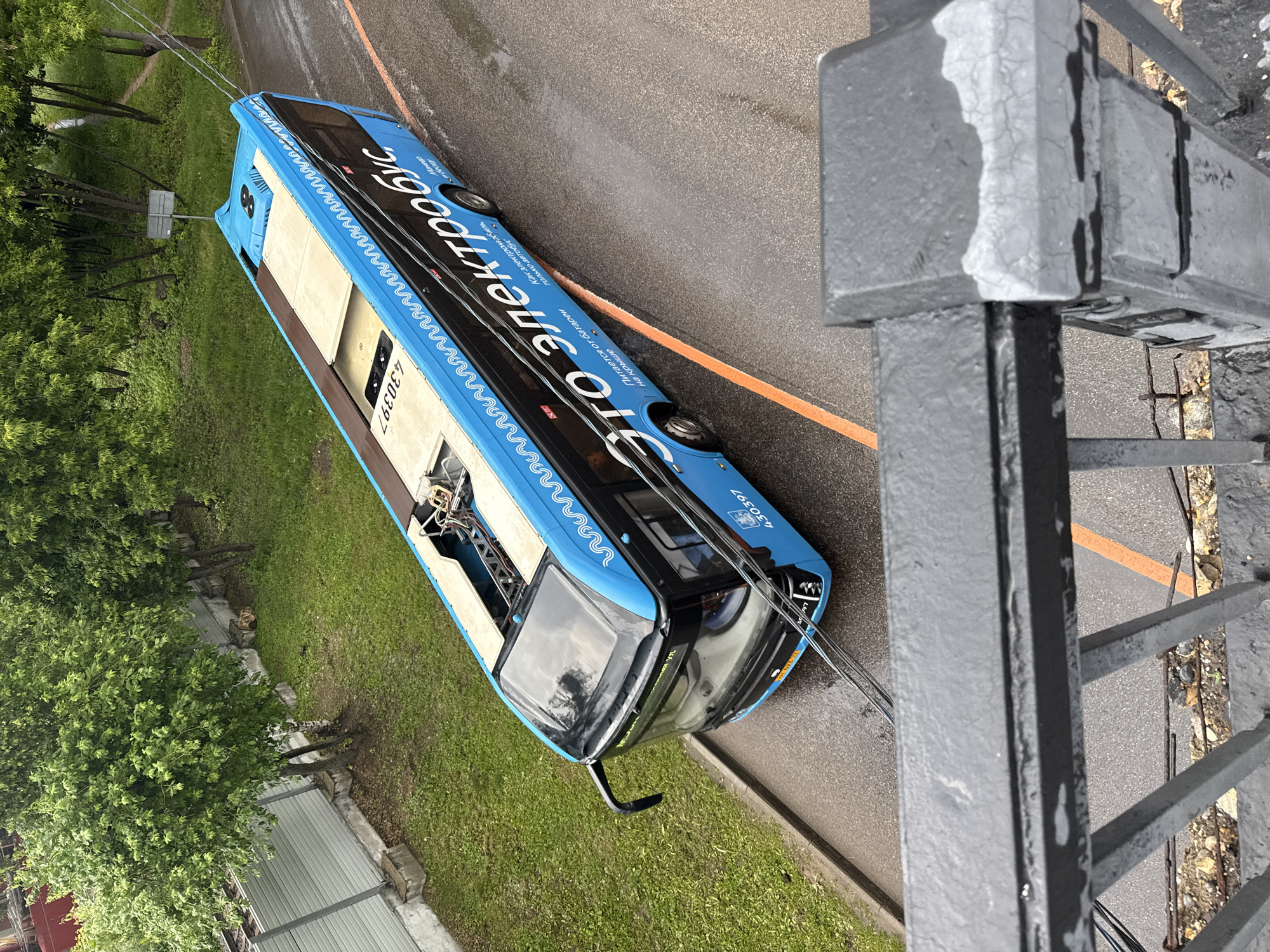 An overhead shot captures a bright blue electric bus with "ЭТО ЭЛЕКТРОБУС" (This is an electric bus) written in large white letters on its side, driving on a wet asphalt road. The bus has a light-colored roof and visible electrical components on top. A license plate on the front of the bus reads "А560ЕР 177 RUS". The bus is on a road with orange lane markings. To the left of the road is a grassy area with trees and a light-colored fence. In the foreground, a dark gray metal railing is visible, suggesting the photo was taken from a higher vantage point, possibly a bridge or building. The overall impression is a city street scene in Moscow, Russia, on a damp day.