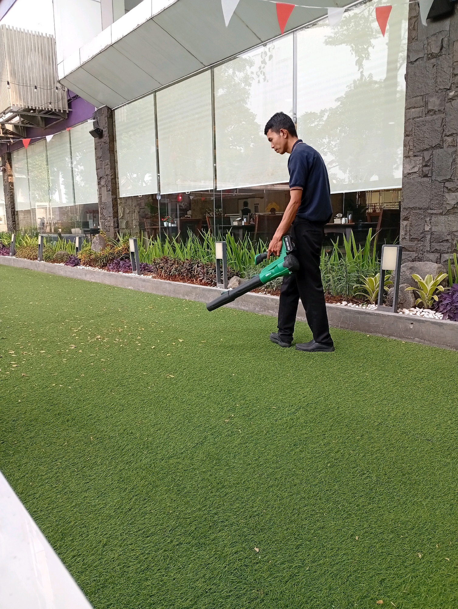 A man in a dark blue shirt and black pants is standing on a bright green artificial turf lawn, holding a green leaf blower. He is facing to his right and looking down. The leaf blower is pointed towards the ground in front of him. Behind him is a long planter box filled with various green and purple plants, separated from the lawn by a low concrete border. Beyond the plants, there is a building with large windows covered by white roller blinds. The building has a dark stone facade and a white overhang. Small red and white triangular flags are strung across the top of the overhang. The scene appears to be outdoors, possibly in an urban area in Binong, Indonesia, given the style of the building and landscaping.
