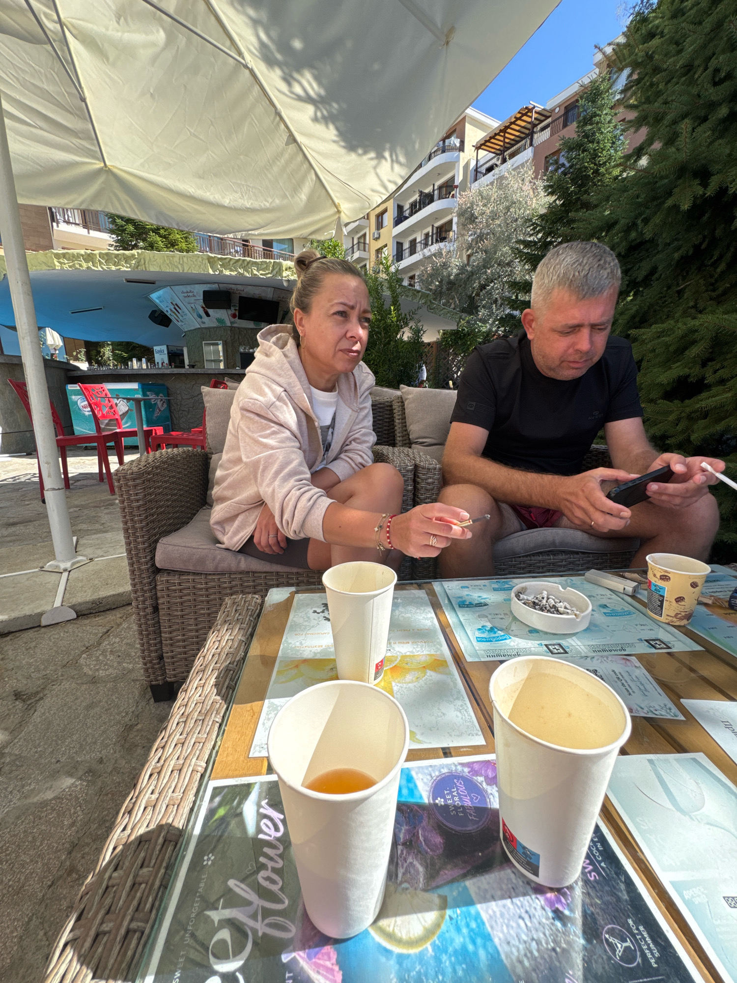 Two adults are seated on a wicker couch under a large white umbrella, outdoors. A man in a black t-shirt and red shorts is on the right, looking at his phone and holding a cigarette. A woman with her hair in a bun, wearing a light pink hooded jacket and dark shorts, is on the left, holding a cigarette and looking to her right. In front of them is a wicker table with several paper cups, one containing a light brown liquid, an ashtray with cigarette butts, and menus or placemats. In the background, a building with multiple balconies and trees are visible, along with a bar area with red chairs and a cooler. The location appears to be a cafe or restaurant in Sveti Vlas, Bulgaria.