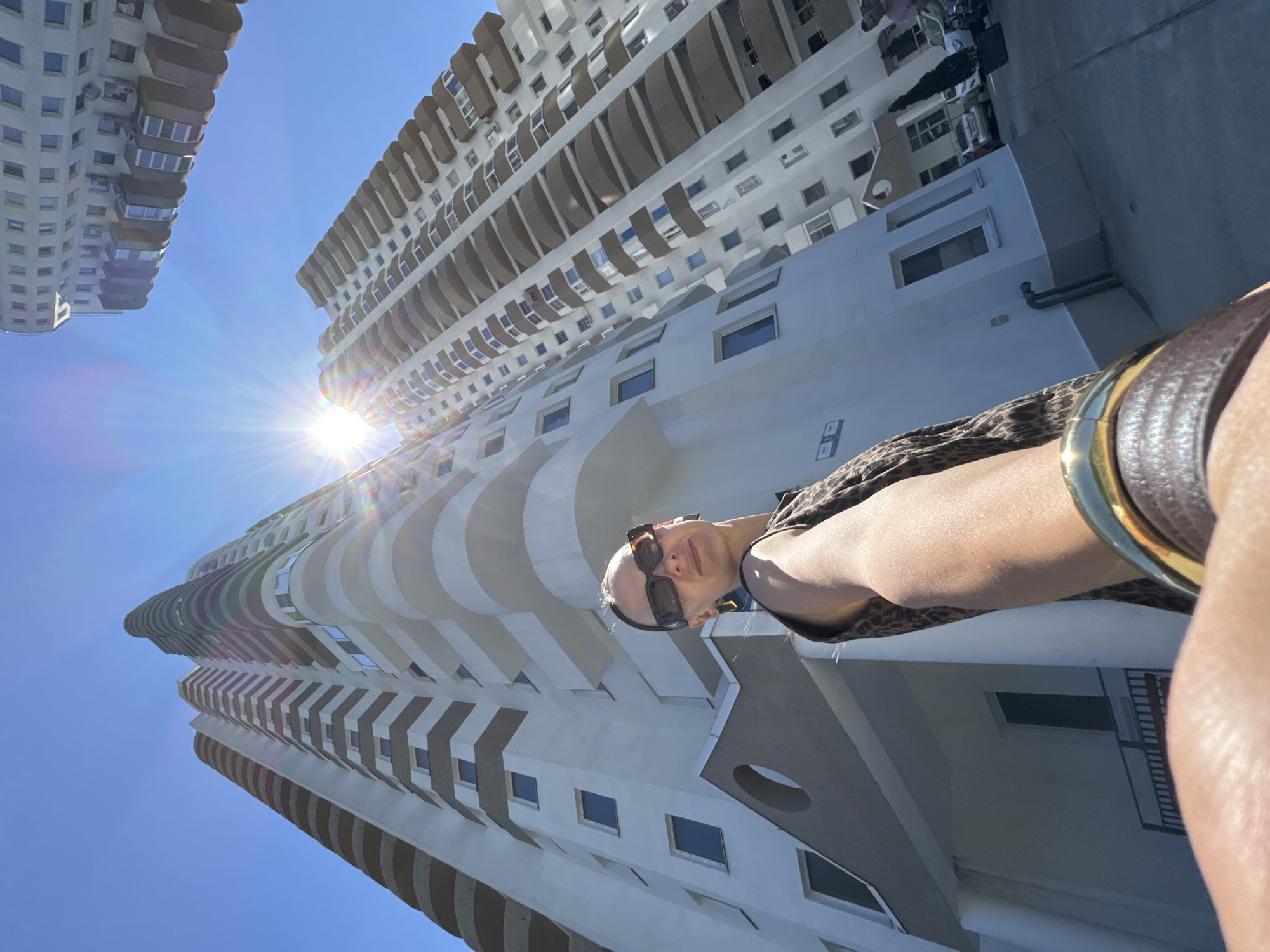 The image is a low-angle shot, taken outdoors on a sunny day, possibly in Sochi, Russia. The focus is on a modern, tall building with numerous curved balconies and many windows. The building dominates the frame, stretching upwards towards a clear blue sky. Sunlight beams brightly, creating lens flares and illuminating the architecture. In the foreground, a person is partially visible, holding the camera for a selfie. They are wearing sunglasses and a leopard-print top, and a large bracelet can be seen on their wrist. The perspective makes the building seem overwhelming and grand, with the person appearing small in comparison. The overall mood is bright and summery, suggesting a leisurely day. No text is visible in the image.