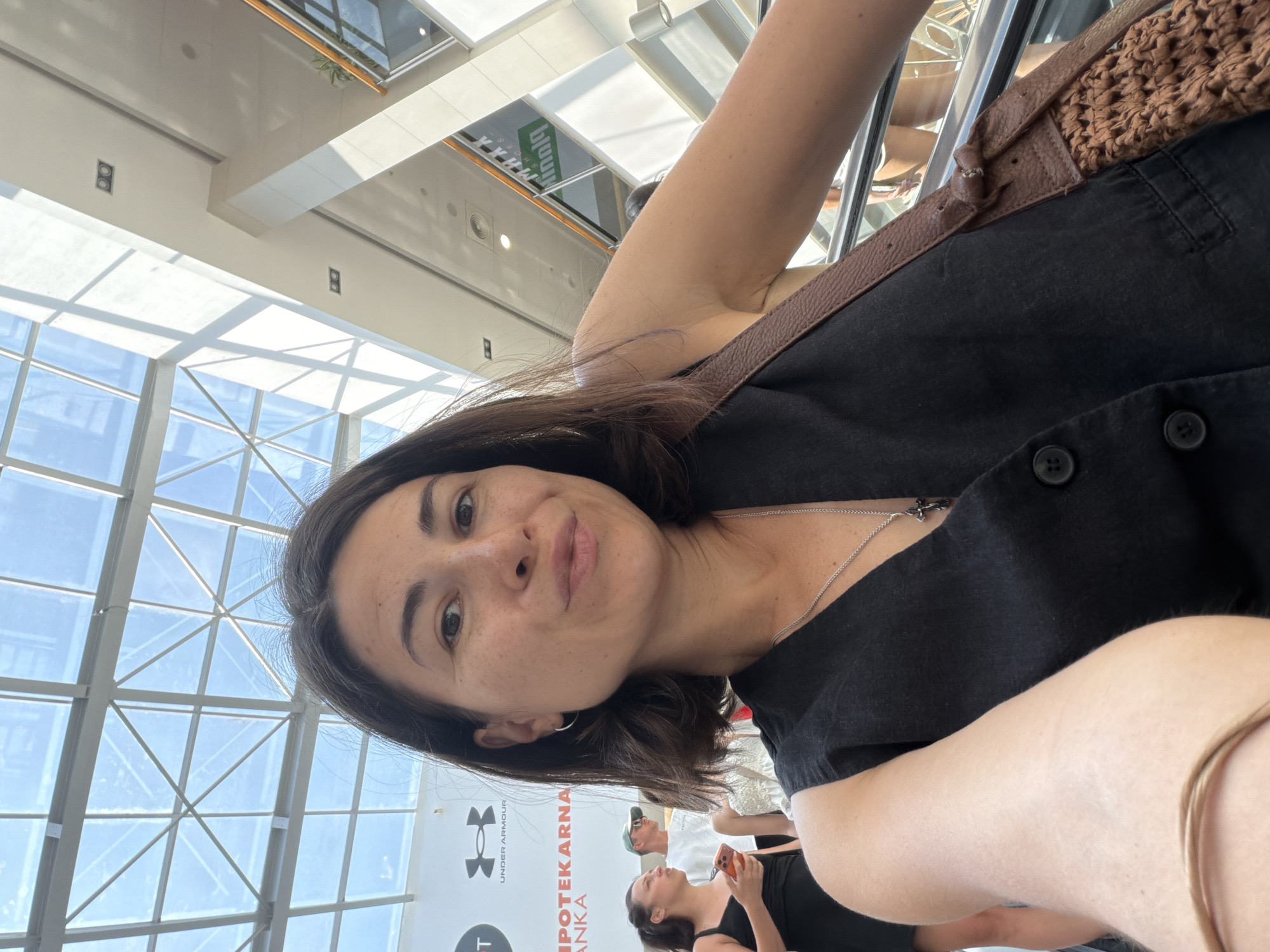The image is a selfie taken by a woman inside a modern shopping mall with a glass ceiling. The woman is wearing a black sleeveless vest over a silver necklace with a cross pendant, and a brown woven purse is slung over her shoulder. She is looking at the camera with a slight pout. In the background, other shoppers can be seen, including a woman in a black dress and a man wearing a white t-shirt and a cap. A large banner advertising "Under Armour" and "Apotekarna Janka" is visible on the left side of the image. The mall appears to be bright and airy, with natural light streaming in from the ceiling. The location is identified as Podgorica, Montenegro.