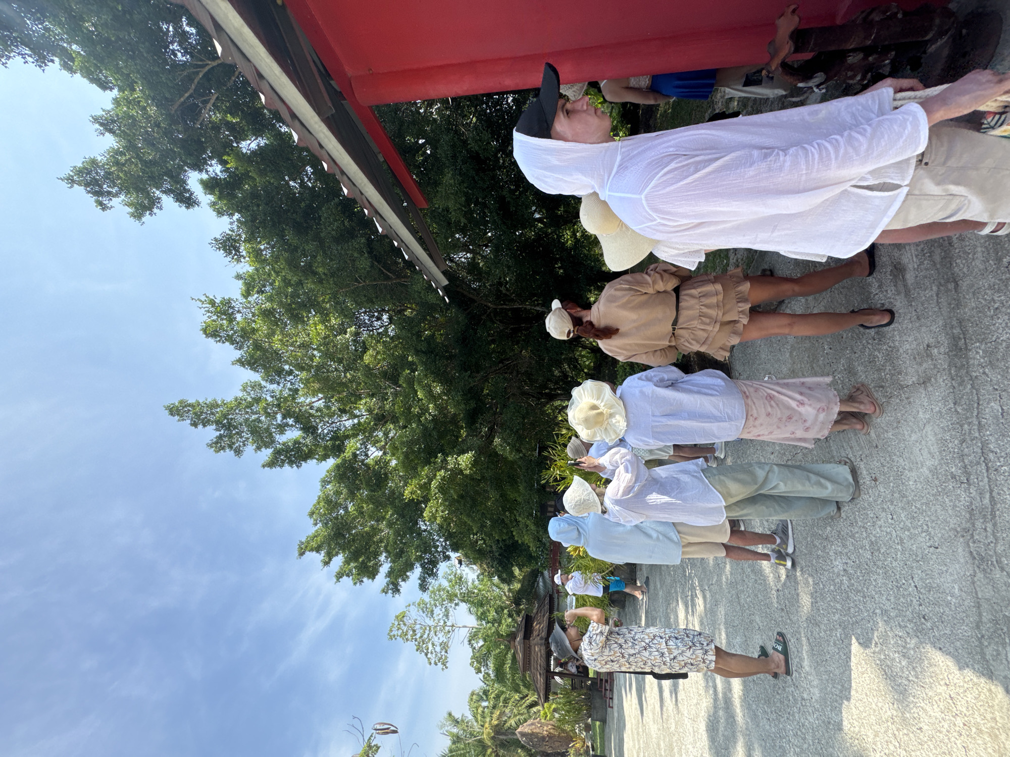 The scene depicts a group of at least seven individuals standing outdoors in Ban Tha Ruea, Thailand. They appear to be interacting near a red-painted structure with a tiled roof, possibly a temple or significant building. The people are dressed in varied casual attire, some wearing sun hats and light-colored clothing. The environment includes trees, vegetation, a paved surface, and a view of a blue sky.