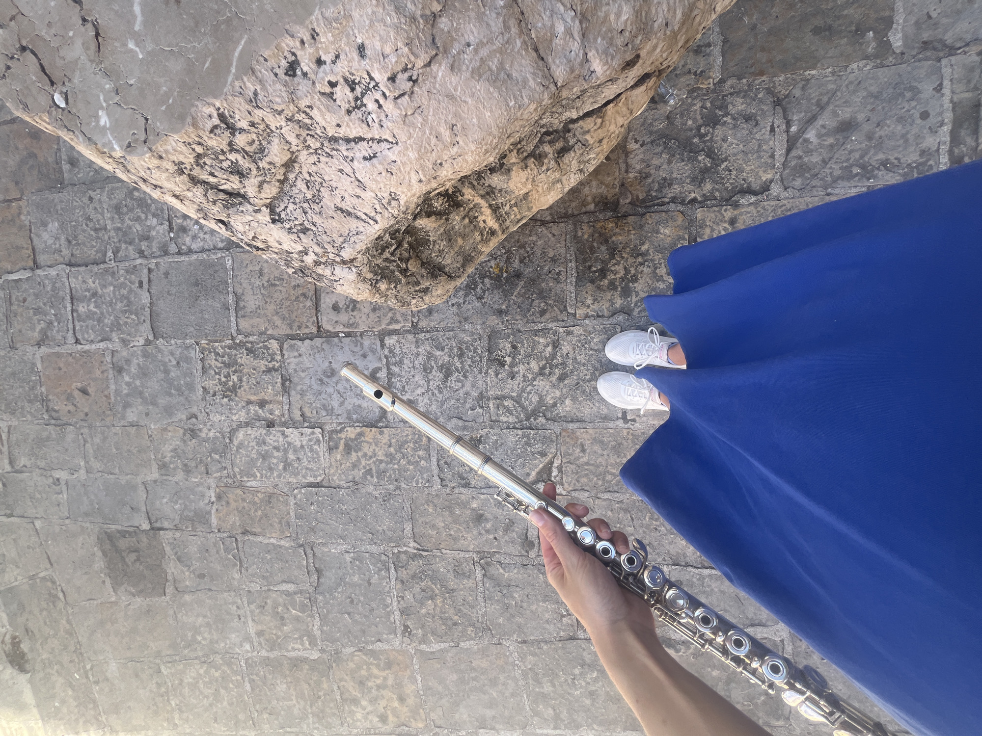The image shows a first-person perspective shot taken outdoors. The background consists of a cobblestone surface, with a large, light-colored stone structure or ruin occupying the upper left corner of the frame. The cobblestones and stone structure suggest a historic location. In the upper right quadrant, there is a section of blue fabric, possibly a dress or tablecloth.
The main subject is a silver flute, held in the lower half of the image by a hand. The flute is angled diagonally, with the mouthpiece pointing upwards. The hand holding the flute appears to be light-skinned.
Near the upper right corner, there is a pair of white sneakers visible. These likely belong to the person taking the photo.
Considering the cobblestone pavement and the presence of the stone structure, it is likely this photo was taken in or around the Old Town area of Budva, Montenegro. The Old Town is characterized by stone architecture, narrow streets, and historic buildings.