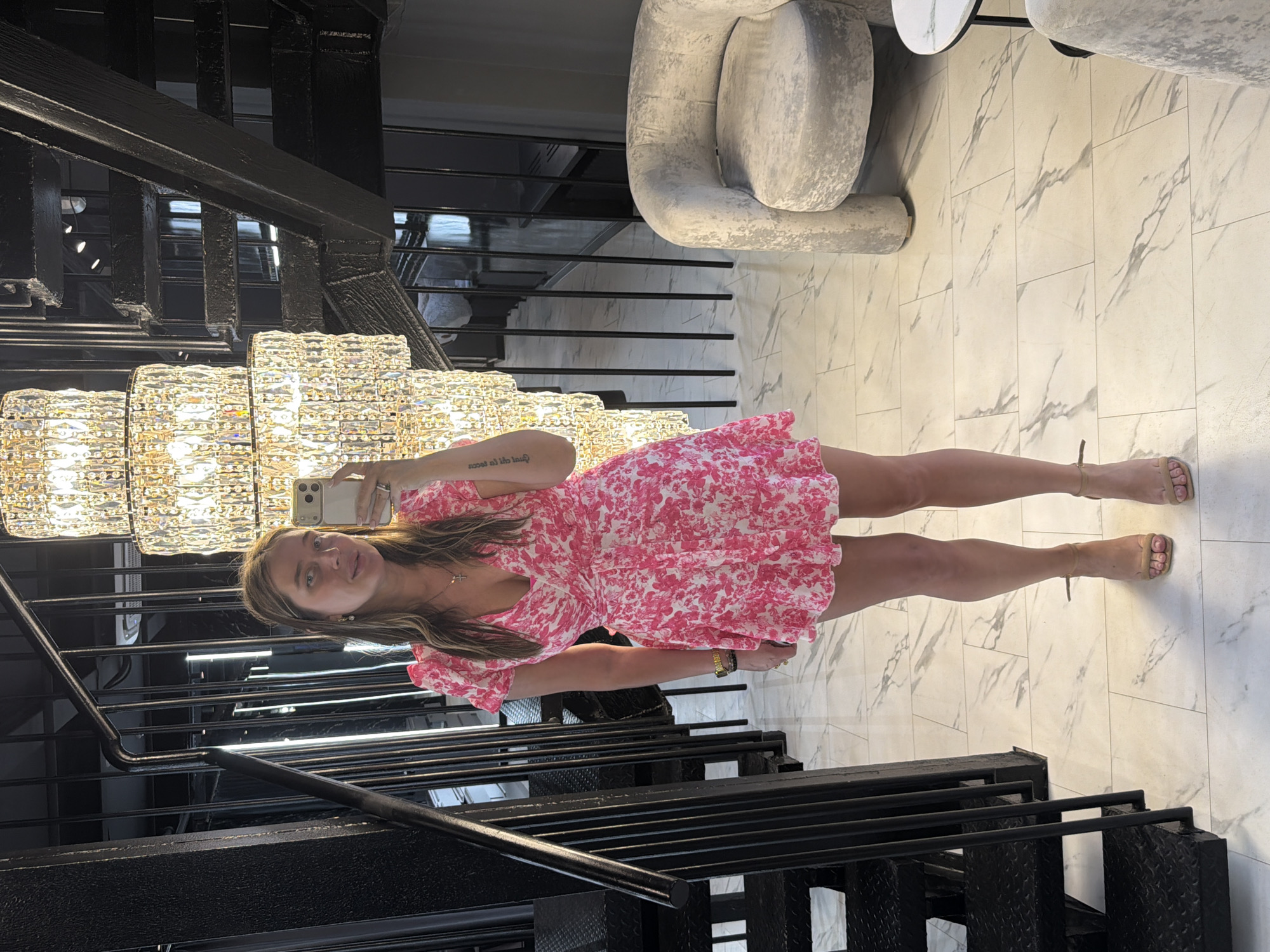 A woman is standing in front of a staircase with a large crystal chandelier overhead. She is wearing a pink floral dress and tan sandals. She is holding a phone in her left hand, taking a selfie. The background includes a gray chair, marble flooring, and black metal railings. The location is potentially a luxurious interior space in Moscow, Russia, due to the ornate design elements and overall upscale atmosphere.