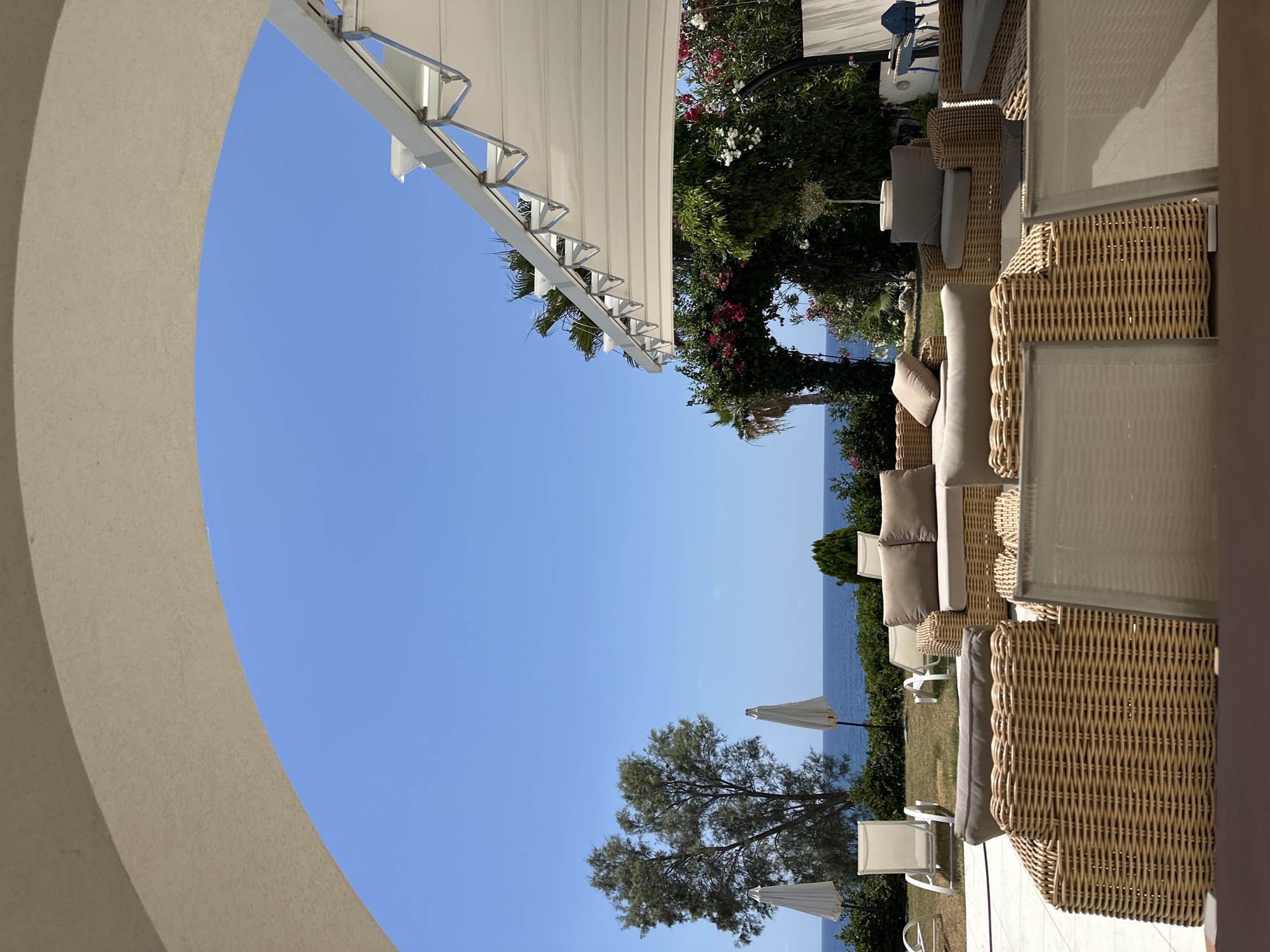 This is a photograph taken outdoors in Kissonerga Community, Cyprus, likely at a hotel or resort, showcasing a lounge area with a view of the sea. The foreground is dominated by the wicker frames of outdoor seating, including sofas and armchairs with beige cushions, arranged on a tiled patio. The middle ground features a manicured lawn with more lounge chairs and closed white umbrellas. Beyond the lawn, the blue sea stretches to the horizon under a clear blue sky. Lush greenery, including trees and flowering bushes, forms a backdrop, with an archway draped in vibrant pink bougainvillea adding a decorative element. A white retractable awning is visible on the right, extending over part of the seating area. The overall impression is one of a serene, sunny, and inviting outdoor space designed for relaxation. There are no people visible in the frame.