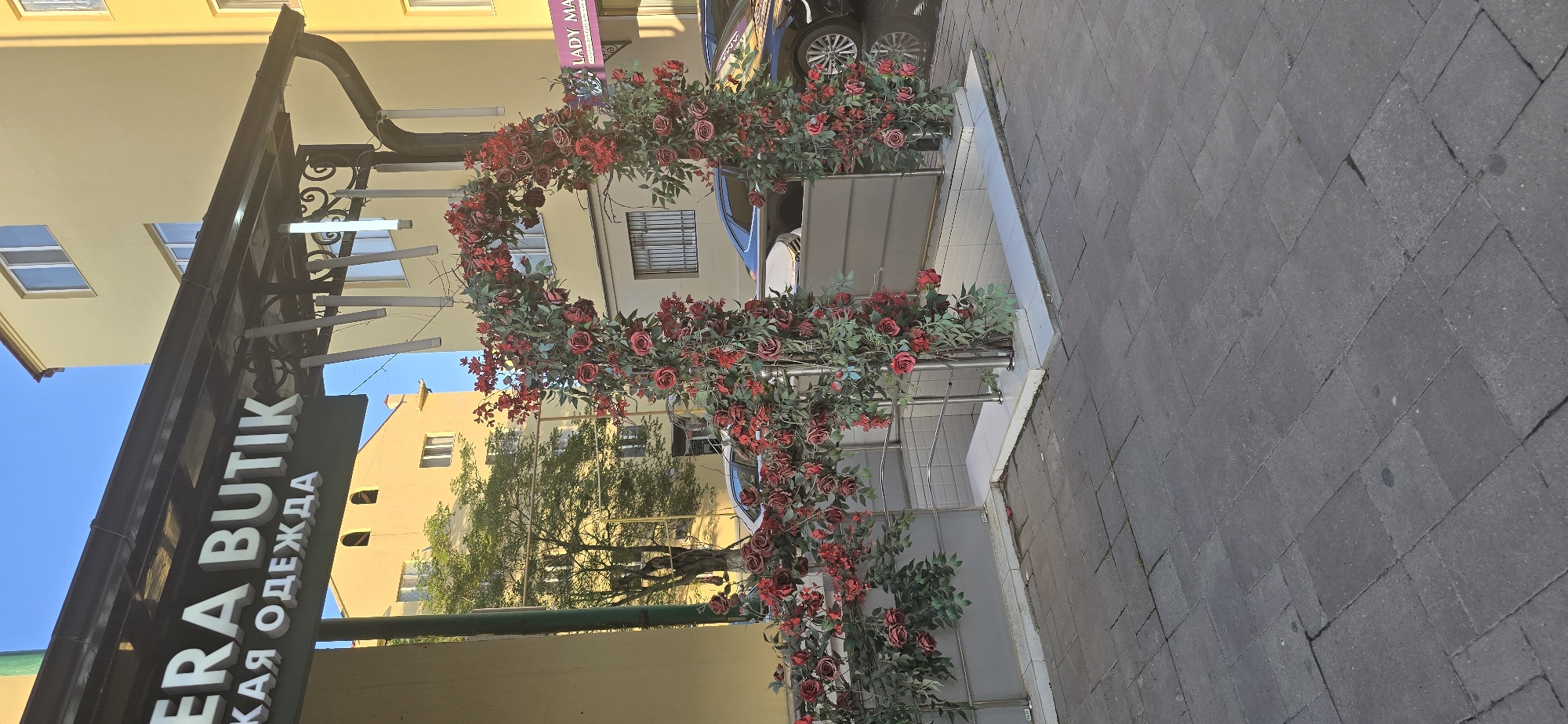 The image shows the storefront of "Era Butik" clothing store in Grozny, Russia. The sign "Era Butik" with "КАЯ ОДЕЖДА" written beneath it, in Cyrillic for "clothing", is visible. An elaborate floral arrangement of red roses adorns a metal archway in front of the store. Nearby buildings, trees and a parked car provide the backdrop. The street is paved with grey brick.