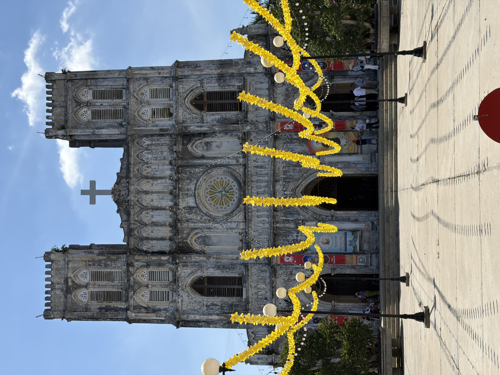 A wide-angle, eye-level, outdoor, daytime photograph captures the facade of a weathered, gothic-style church, likely located in Hội Tín, Vietnam. The church's stone structure is gray and shows signs of age, with two prominent towers flanking the central structure. A large gray cross sits atop the church's apex.
The entrance to the church is recessed, with a wide set of stone stairs leading up to it. The facade is decorated with elaborate architectural details, including arched windows and a large rose window above the entrance. Yellow garlands, made of paper or fabric, are draped across the front of the church, strung between lampposts. Small white string lights are also visible within the garlands. Several banners hang on the church facade, featuring text and images.
In front of the church, on a paved courtyard, several people are gathered. Some are standing near the entrance, while others are seated on a stone bench or standing near trees to the left and right of the frame. Two lampposts with multiple globe lights stand prominently in the foreground, casting long, linear shadows across the courtyard. The sky is blue with scattered white clouds.