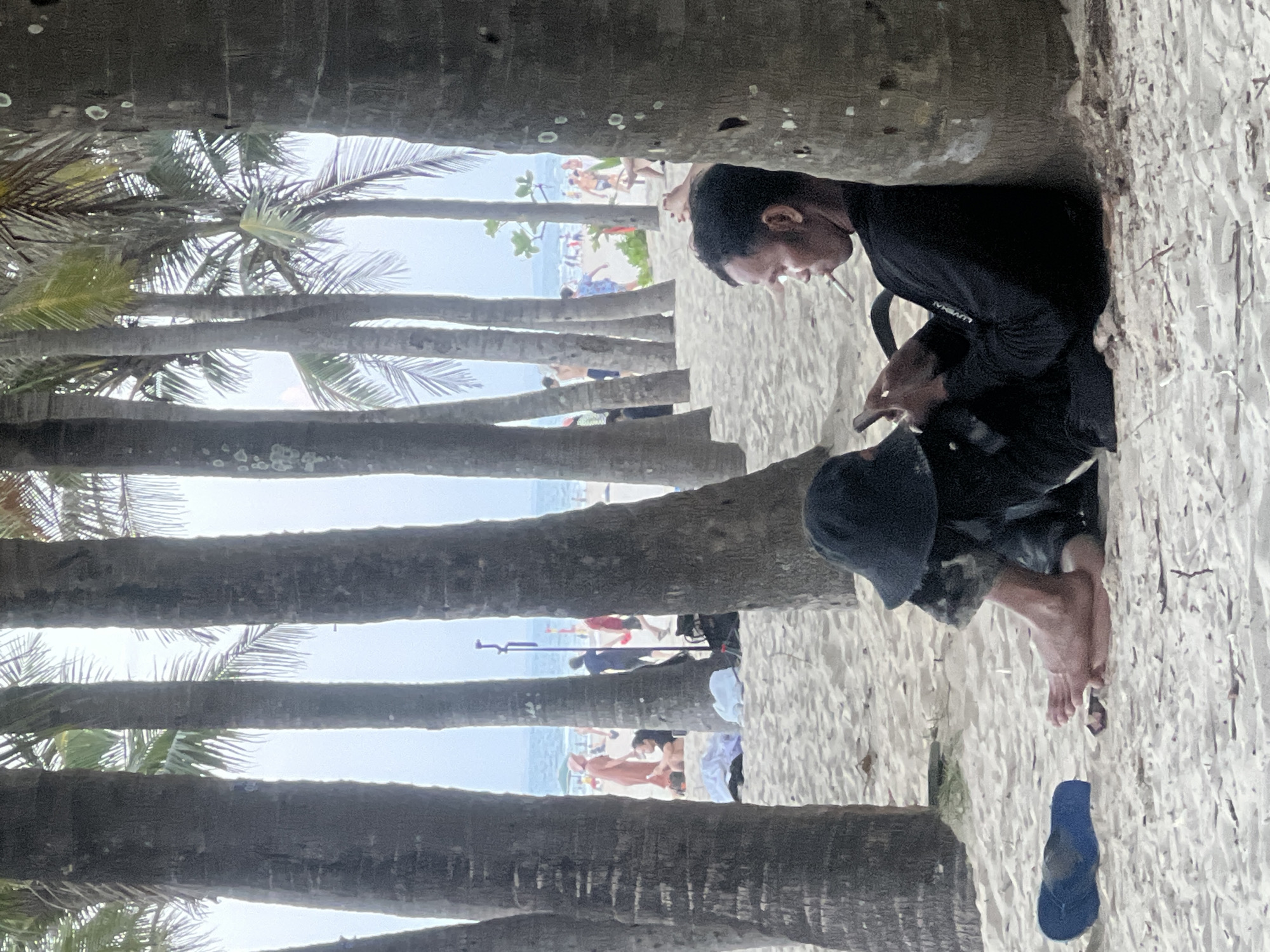 In Đà Nẵng, Vietnam, a man sits on a sandy beach, leaning against the trunk of a palm tree. He wears a black long-sleeved shirt and dark pants, with a black bucket hat covering his head. His legs are crossed, and his feet are bare, resting on the sand. A blue flip-flop lies on the sand to his left. The man is holding a smartphone in his right hand and a cigarette in his mouth, actively smoking. He appears to be looking at the phone, engrossed in its screen. Behind him, other beachgoers are visible near the water, some walking and others relaxing. Palm trees are abundant in the foreground, creating a dappled shade over the beach. The ocean is visible in the background.