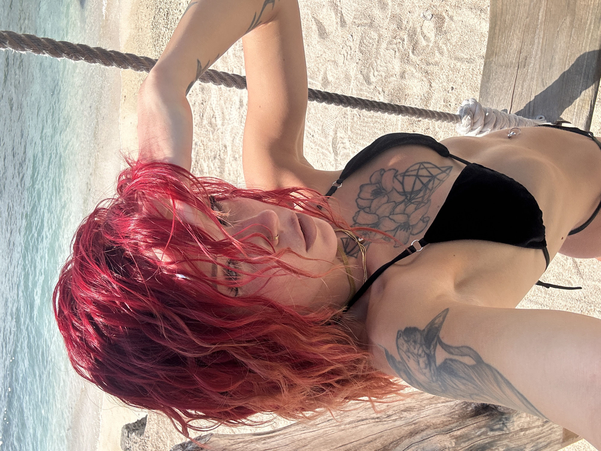 A light-skinned woman with red hair is lying down outside. She is wearing a black bikini. She has multiple tattoos on her arms, chest, and stomach. The woman is lying on a wooden platform. A rope is attached to the wood. The area includes a beach, sand, and water. The geographical location is the city of Unknown, Indonesia.