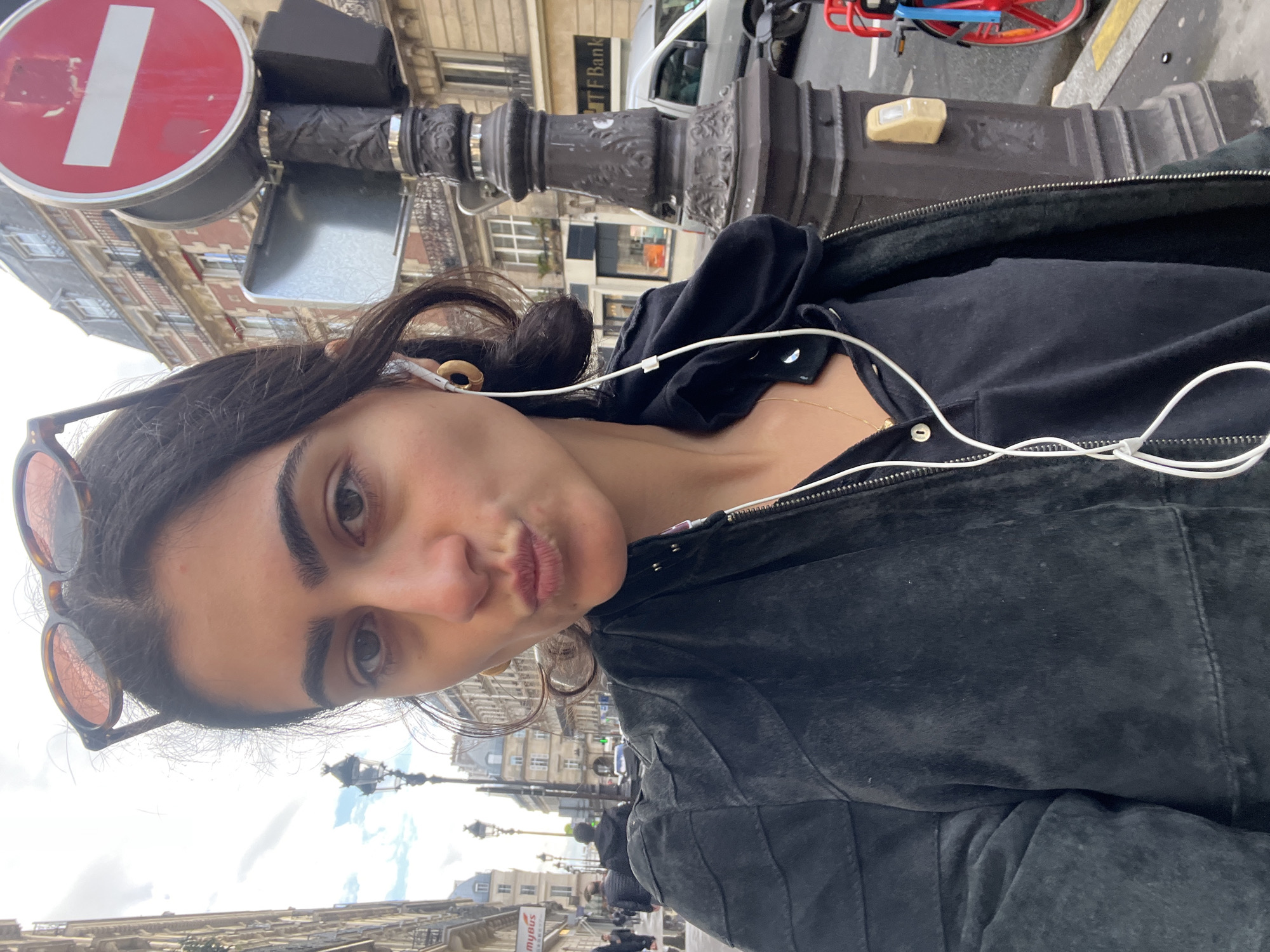 A woman takes a selfie on a Parisian street. She is wearing a black jacket and sunglasses, and has headphones in her ears. The iconic Parisian architecture is visible in the background, including a street sign with a white horizontal bar on a red background, typical of European no-entry signs. The setting appears to be in Paris, France, with the architecture and street signs aligning with typical Parisian street scenes. There is a bank sign “TF Bank” visible in the background.