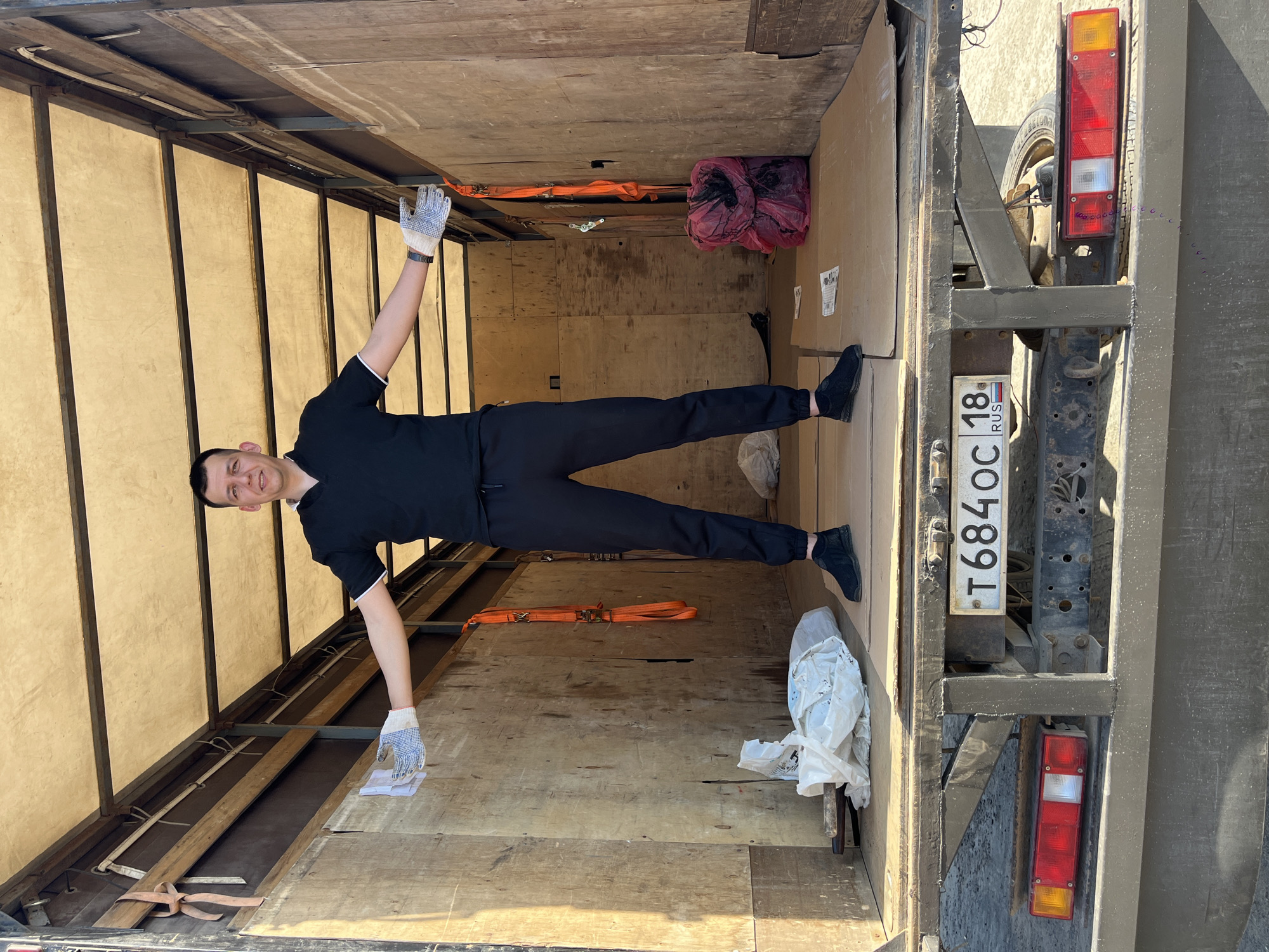 A man in black clothing stands inside a truck, his arms extended outward. He wears work gloves on both hands. The truck's interior is wooden and contains straps, bags, and cardboard boxes. A Russian license plate reading "T684OC 18 RUS" is visible on the back of the truck. The location is likely Izhevsk, Russia, based on the license plate.